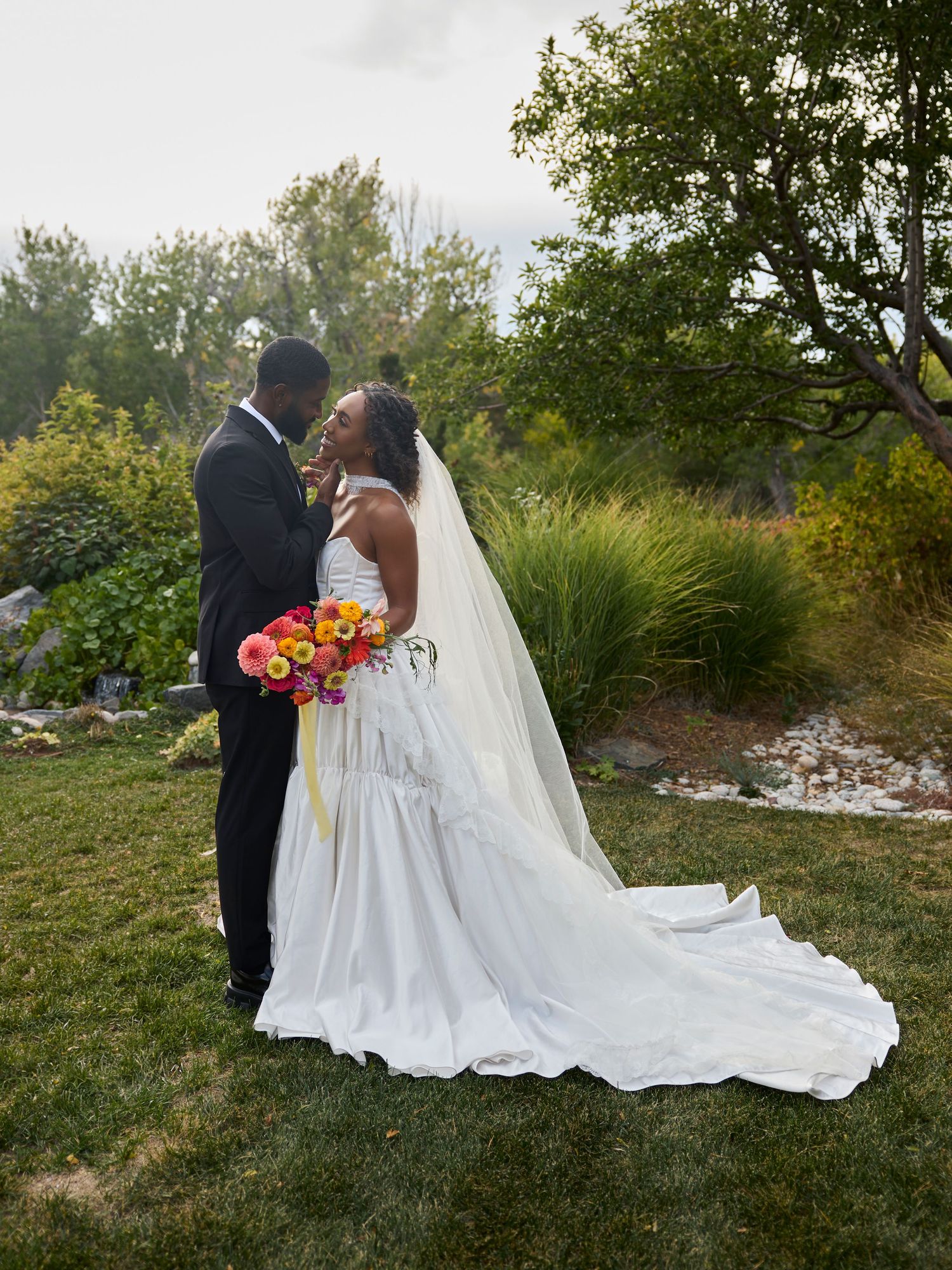 Groom in a black suit kisses the smiling bride in a white gown with a long veil and train in a lush garden.