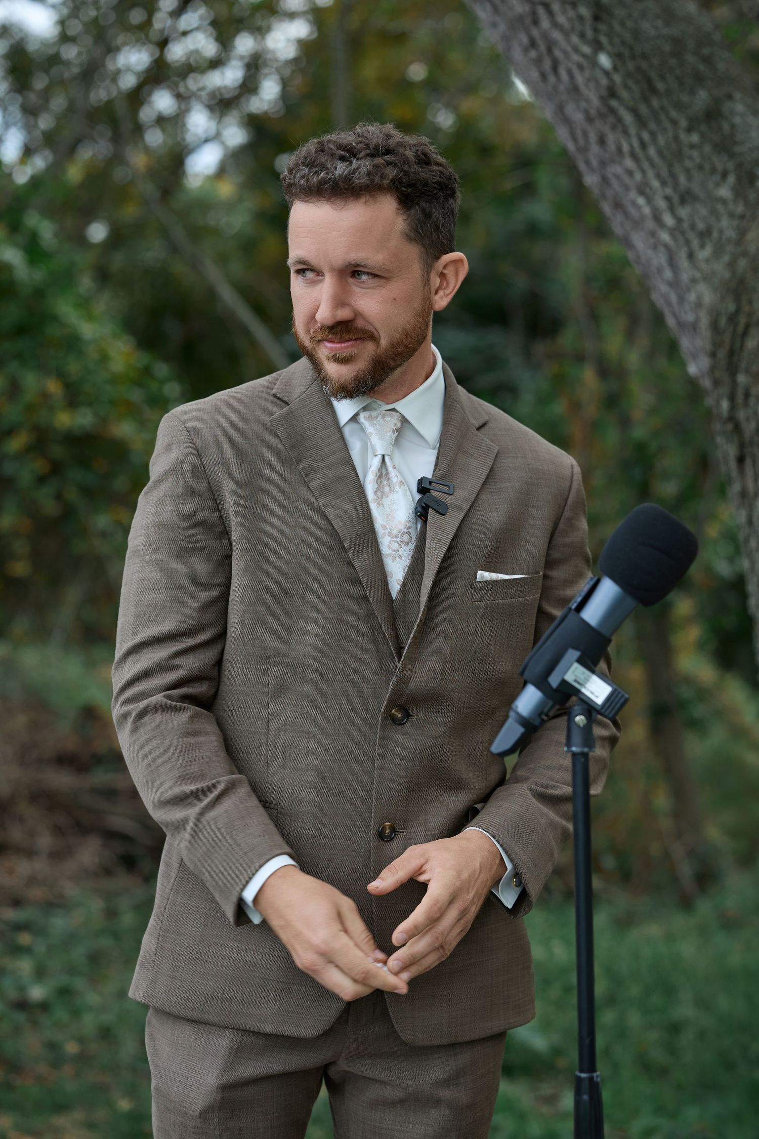Groom in a brown tweed suit with a lapel microphone stands under trees, smiling during his outdoor wedding ceremony.