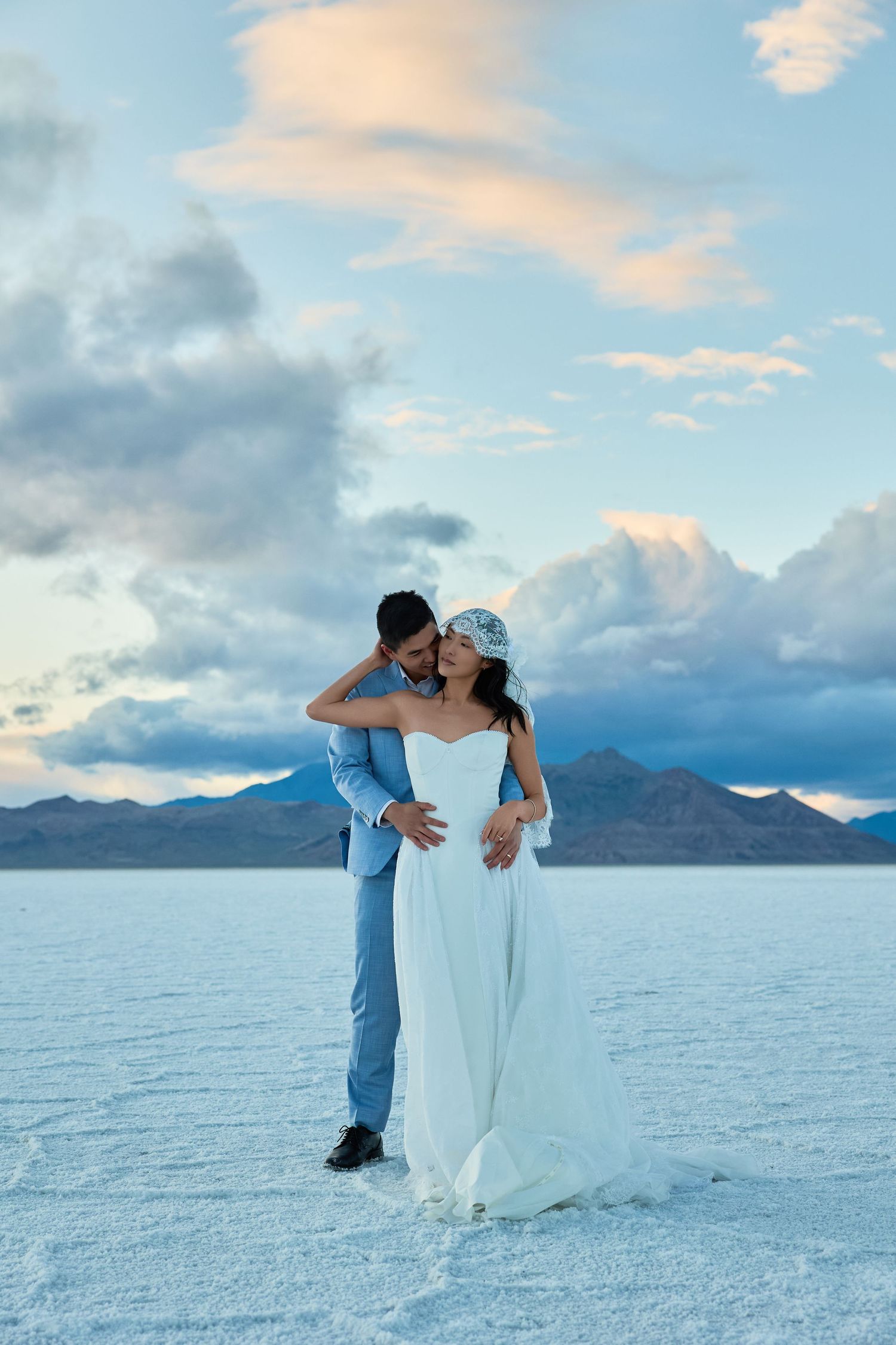 Newlyweds share an intimate embrace on a white salt flat, the bride in a white gown and veil, with mountains and soft clouds beyond.