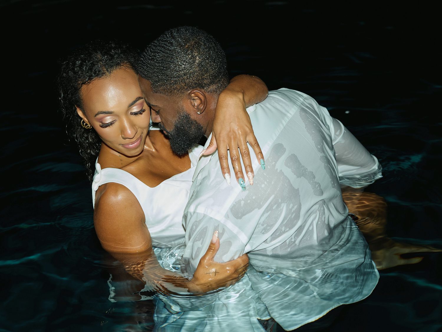 A smiling woman in a white top leans into her partner in a white shirt as they embrace in nighttime pool water.