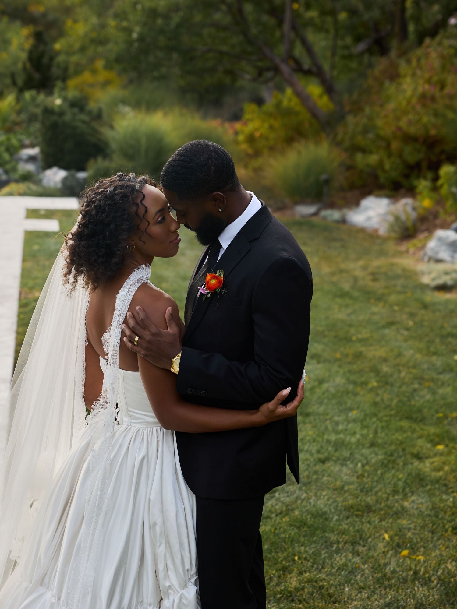 Couple shares a close embrace in a garden, the bride in a veil and white gown, the groom in a black suit with an orange boutonniere.