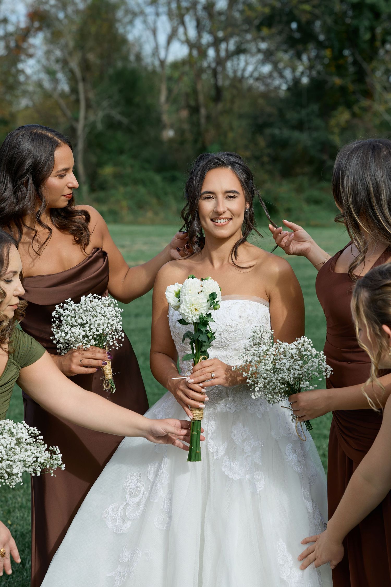 Bride in a lace wedding gown smiles with her bridesmaids in brown dresses holding baby's breath bouquets on a lawn.
