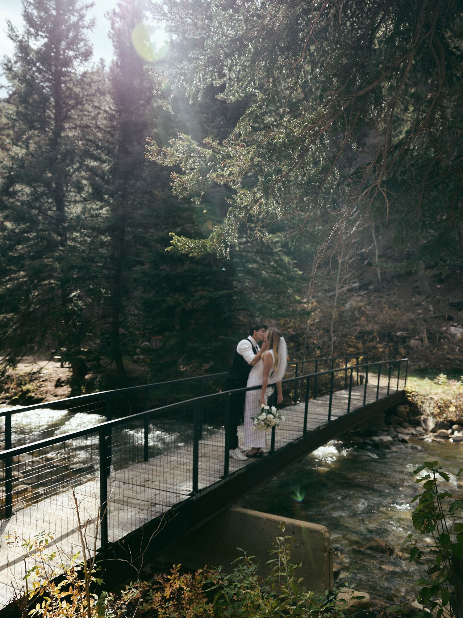 Elopement couple kisses on a metal footbridge over a rocky mountain stream with sunlight filtering through the pine trees.