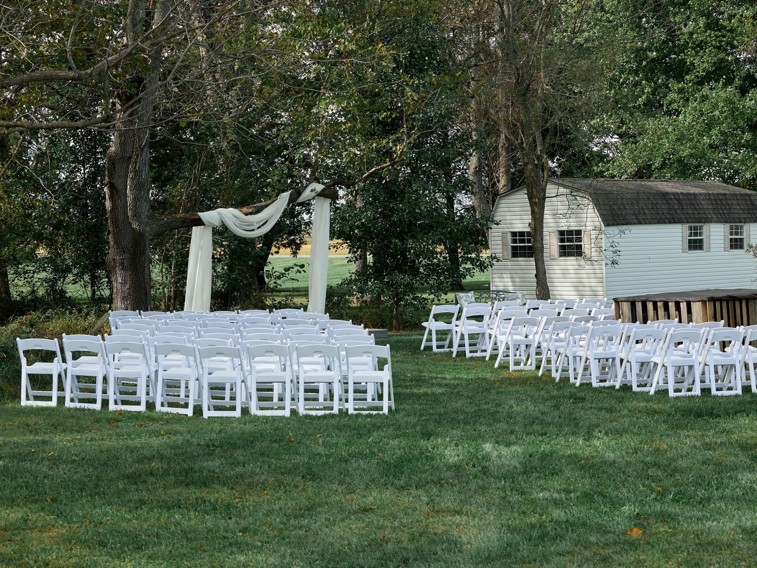 Rows of white folding chairs set up for an outdoor wedding ceremony beneath tall trees with a draped fabric arch.