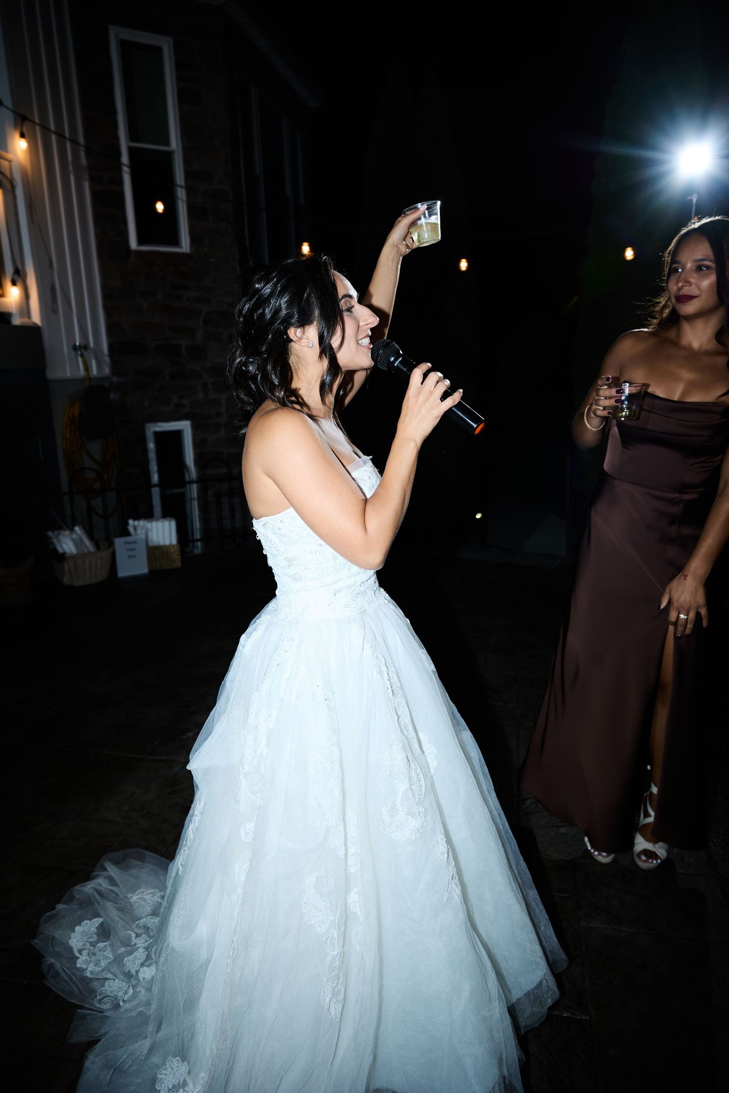 Bride in a white lace gown toasts with a glass and microphone beside a bridesmaid in a brown satin dress at night.