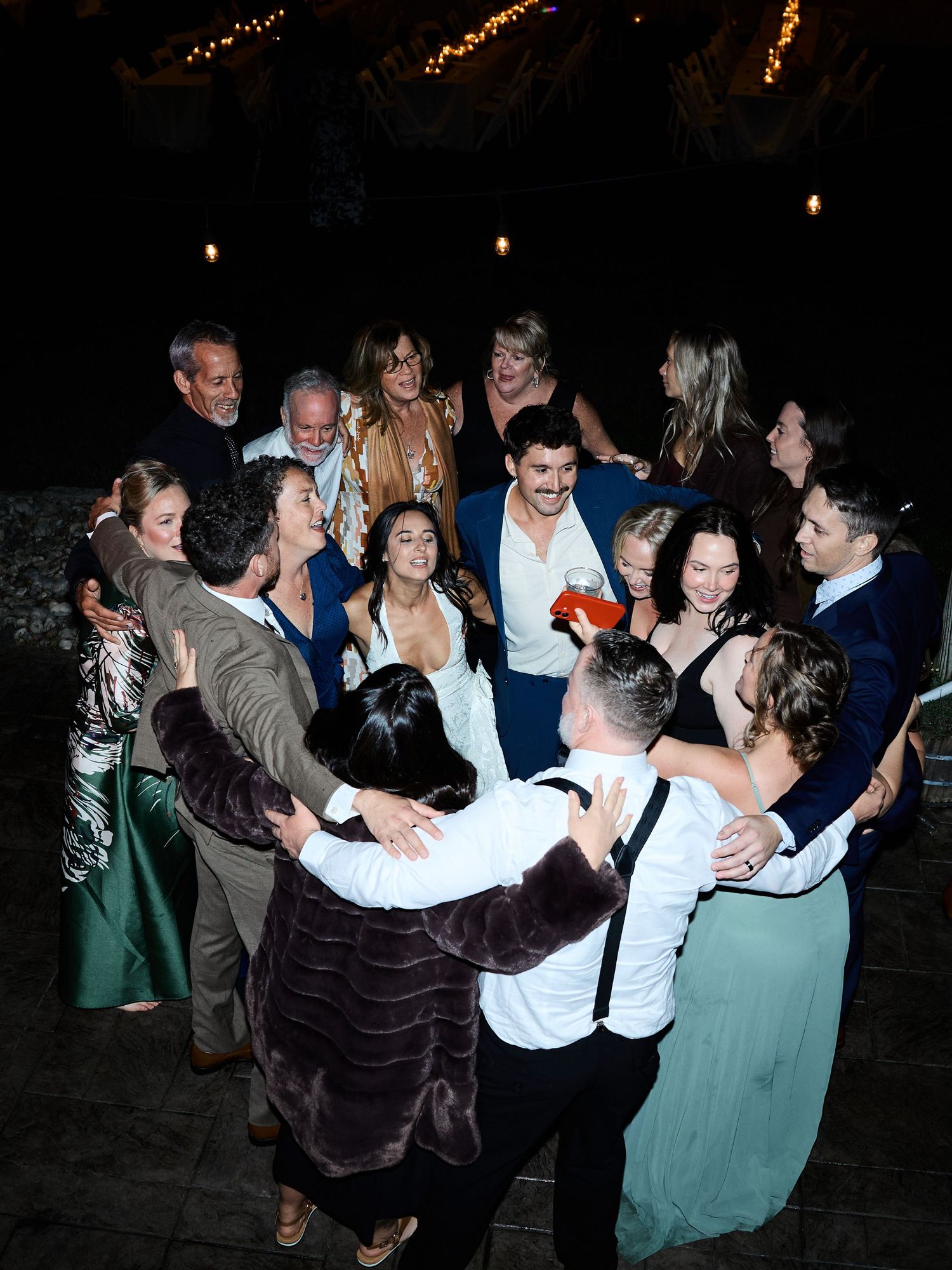 A joyful group of wedding guests in formal attire gather in a tight circle, laughing and dancing together at night.