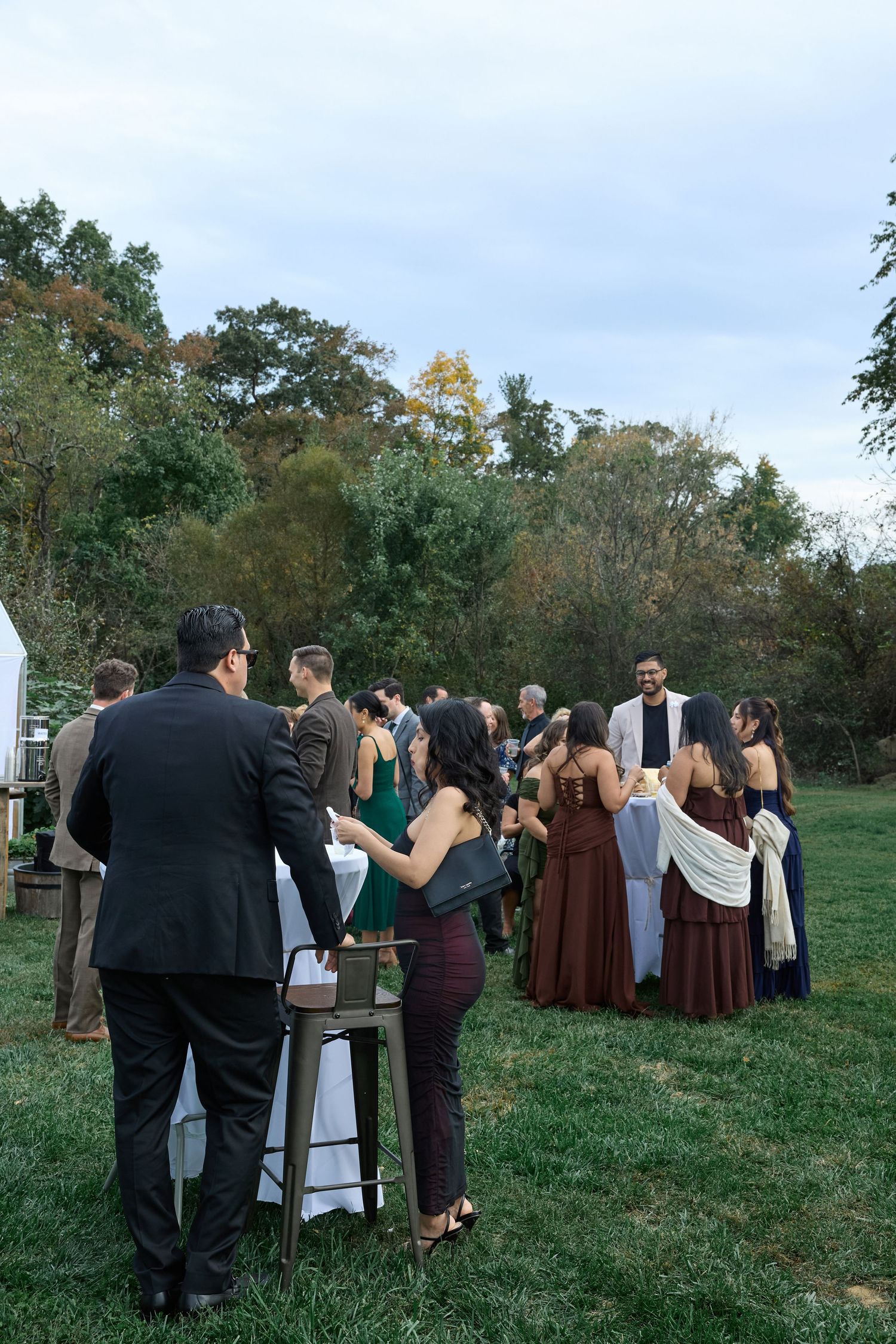 Guests gather in small groups around cocktail tables on a grassy lawn at an outdoor wedding reception with trees in the background.