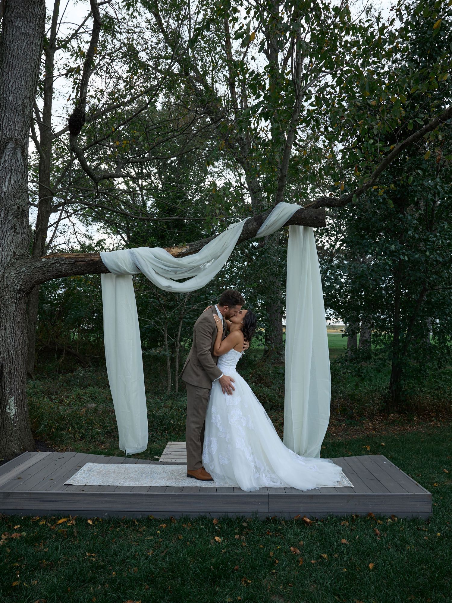 A newlywed couple kisses on a wooden platform beneath a tree draped with flowing white fabric at their outdoor wedding.