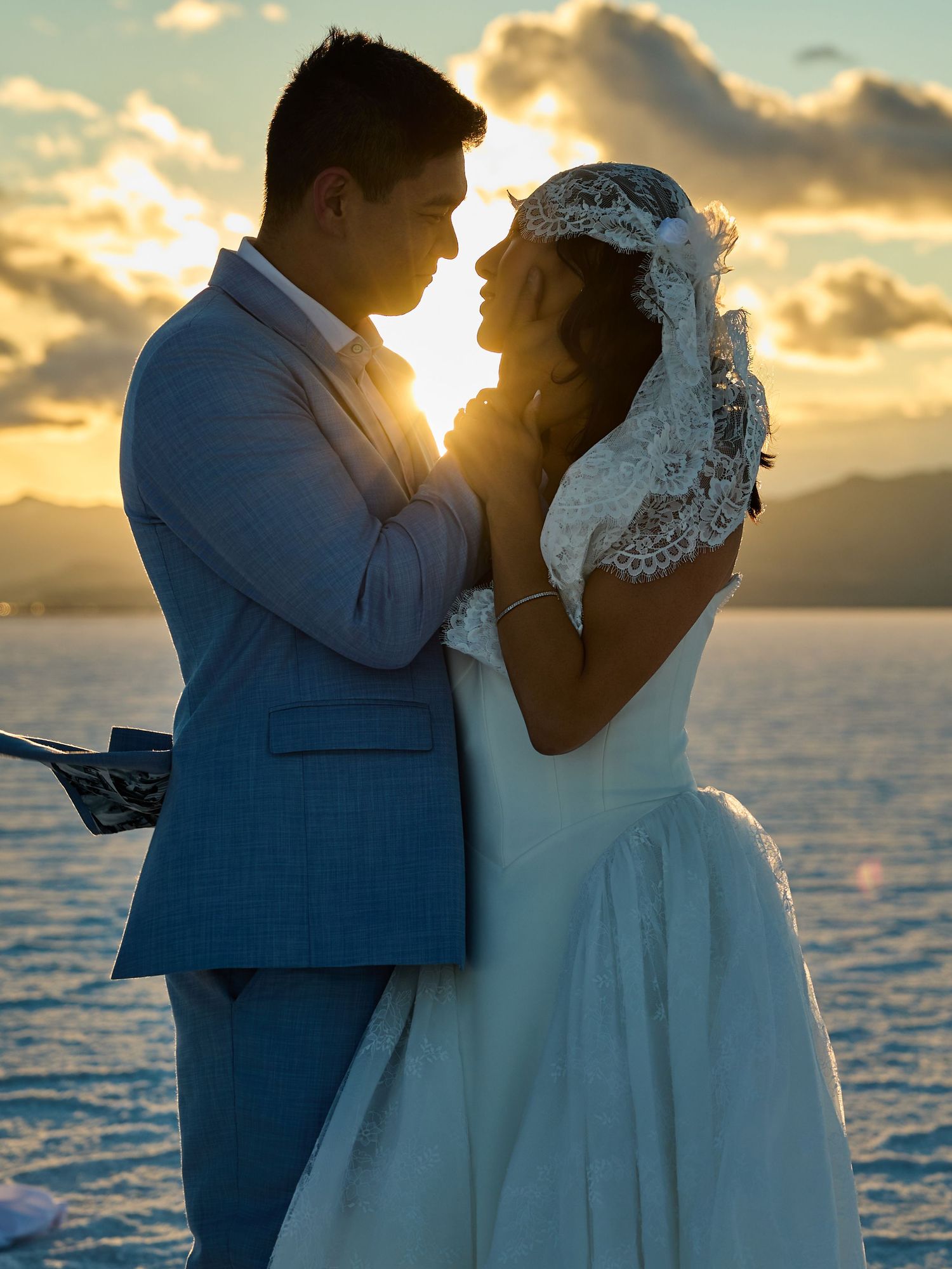 Bride in a lace veil and groom in a blue suit embrace face to face as the golden sunset glows between them over a lake.