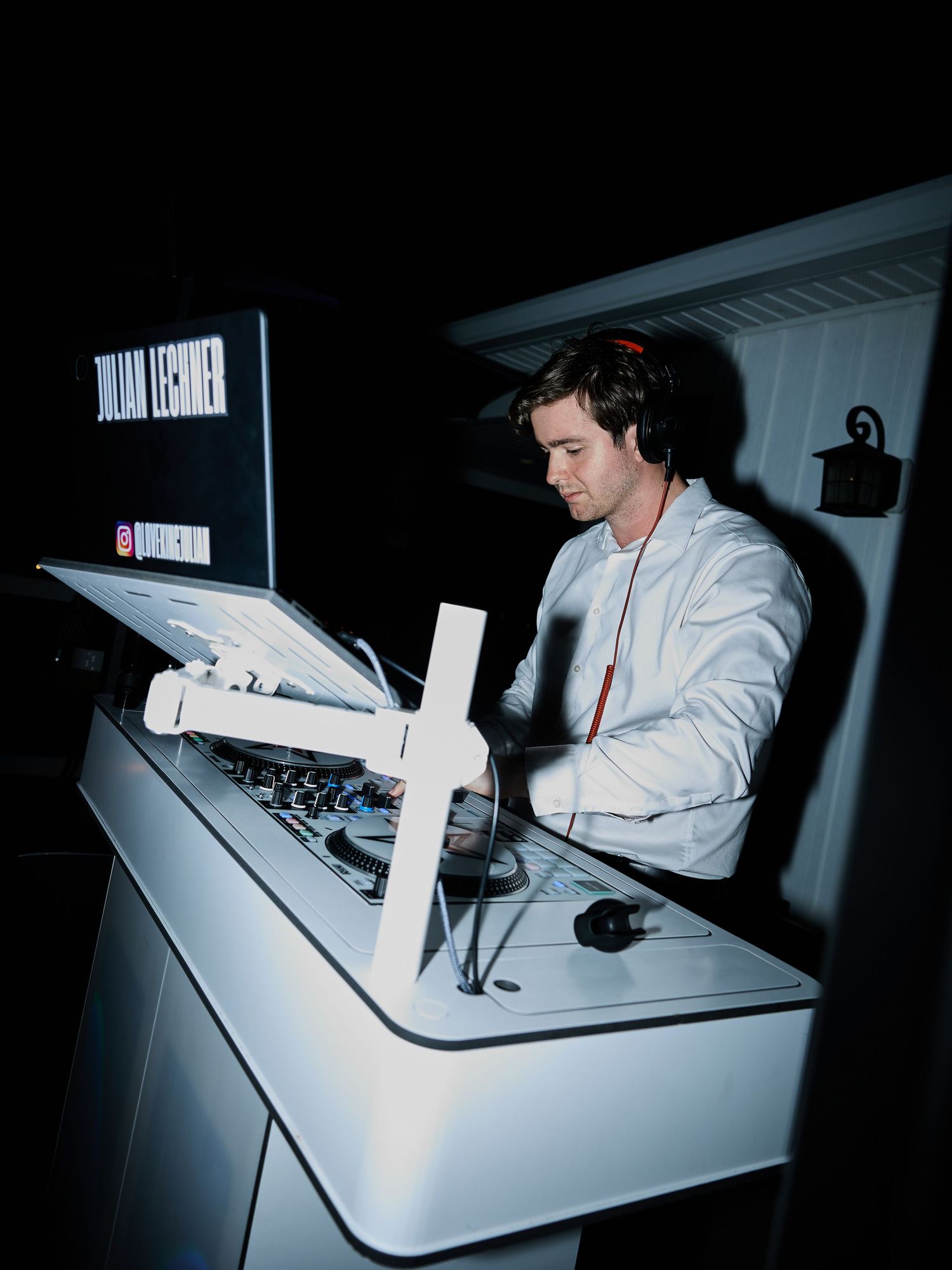 A DJ in a white jacket and headphones performs at a sleek white booth during a nighttime wedding reception.