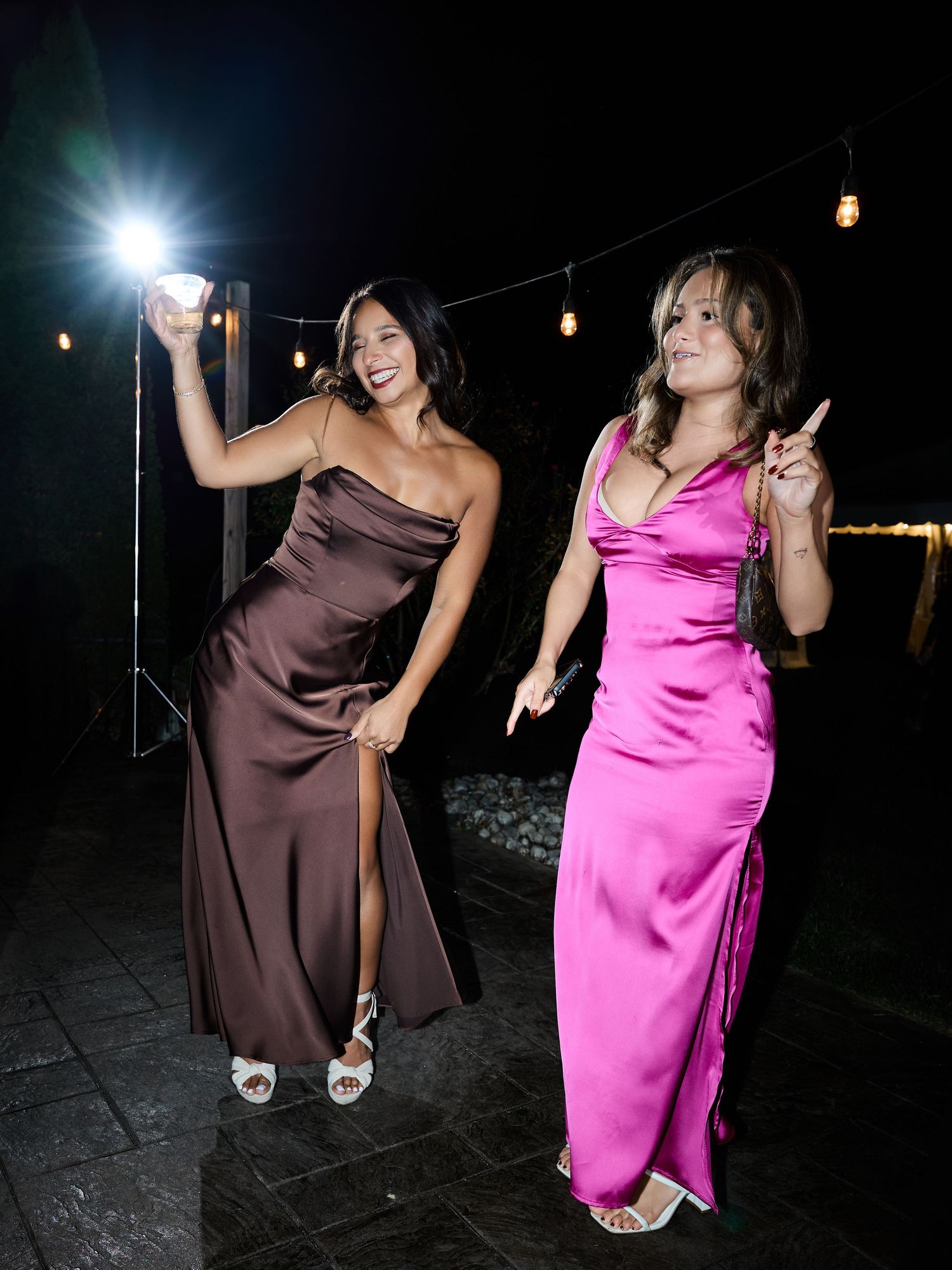 Two women in a brown satin slit dress and a bright pink satin gown dance and smile under string lights at night.