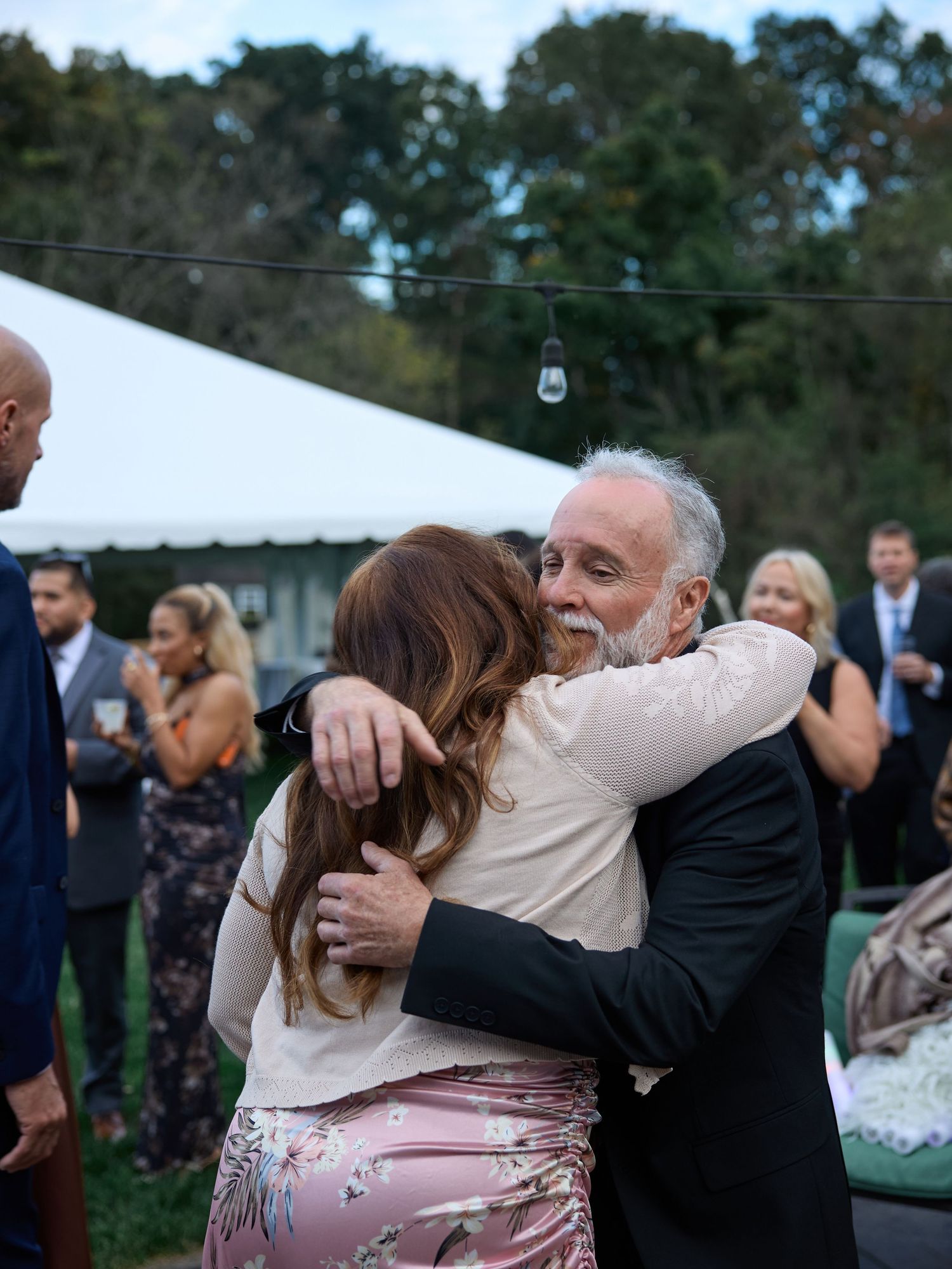 A gray-haired man in a dark suit warmly hugs a woman in a floral skirt at an outdoor wedding reception under string lights.