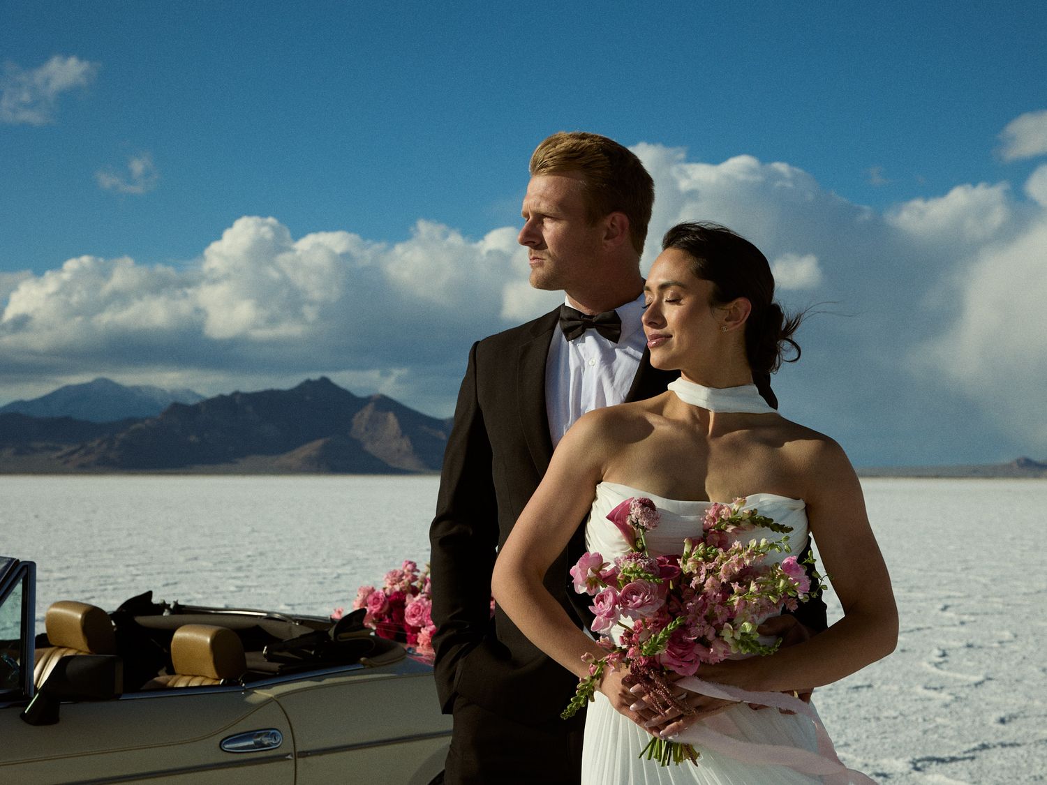 A groom in a tuxedo gazes into the distance as the bride holds a pink bouquet near a vintage car on a salt flat with mountains.