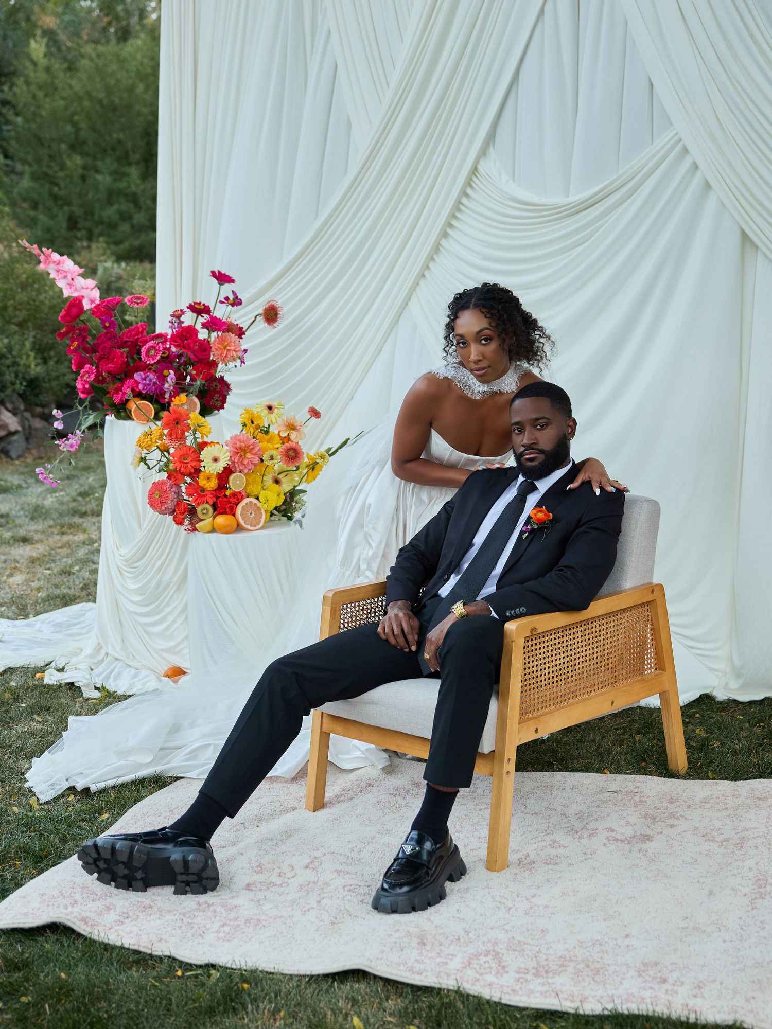 A groom in a black suit sits in a cane chair on a rug while his bride leans over him before draped fabric and flowers.