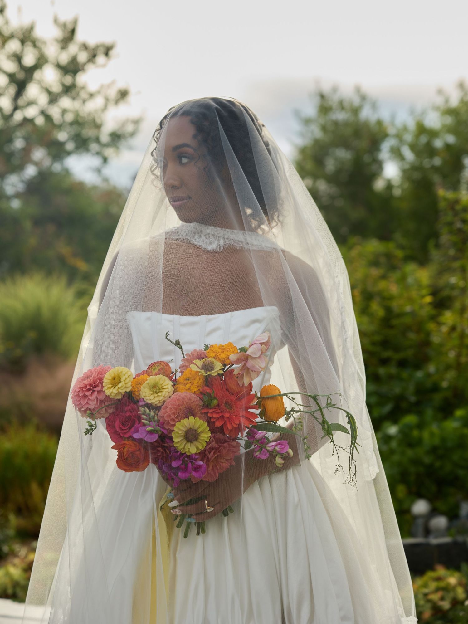 Bride in a strapless white gown and sheer veil holds a vibrant bouquet of orange, pink and yellow flowers outdoors.
