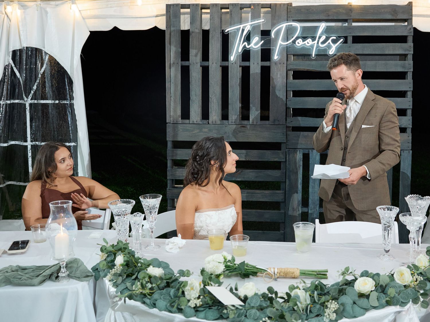 A groom in a brown suit reads from a card into a microphone while the bride sits at a flower-decked head table under a neon sign.