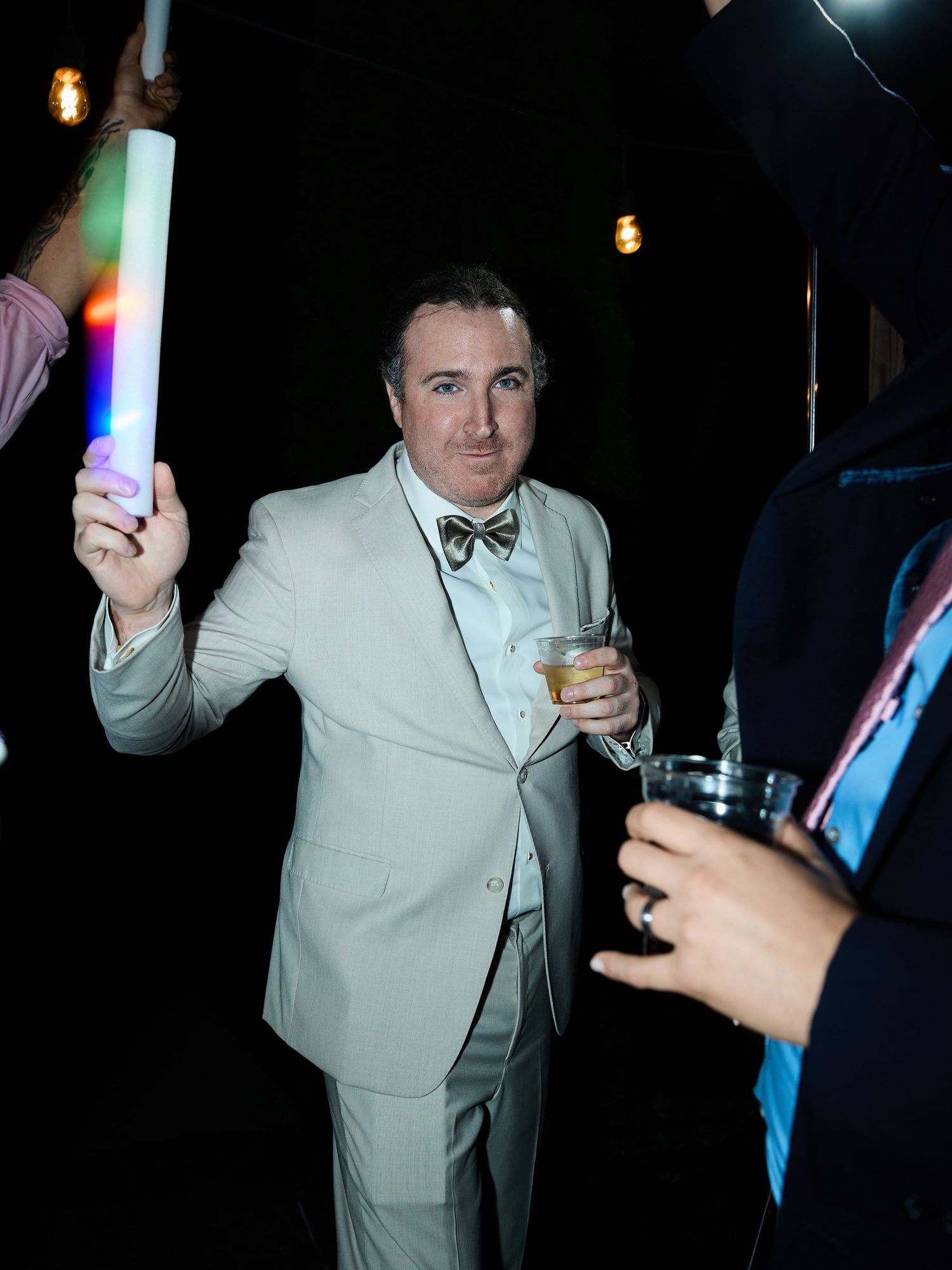 Man in a light gray suit and bow tie holds a drink and a glowing foam stick at a festive nighttime wedding reception.