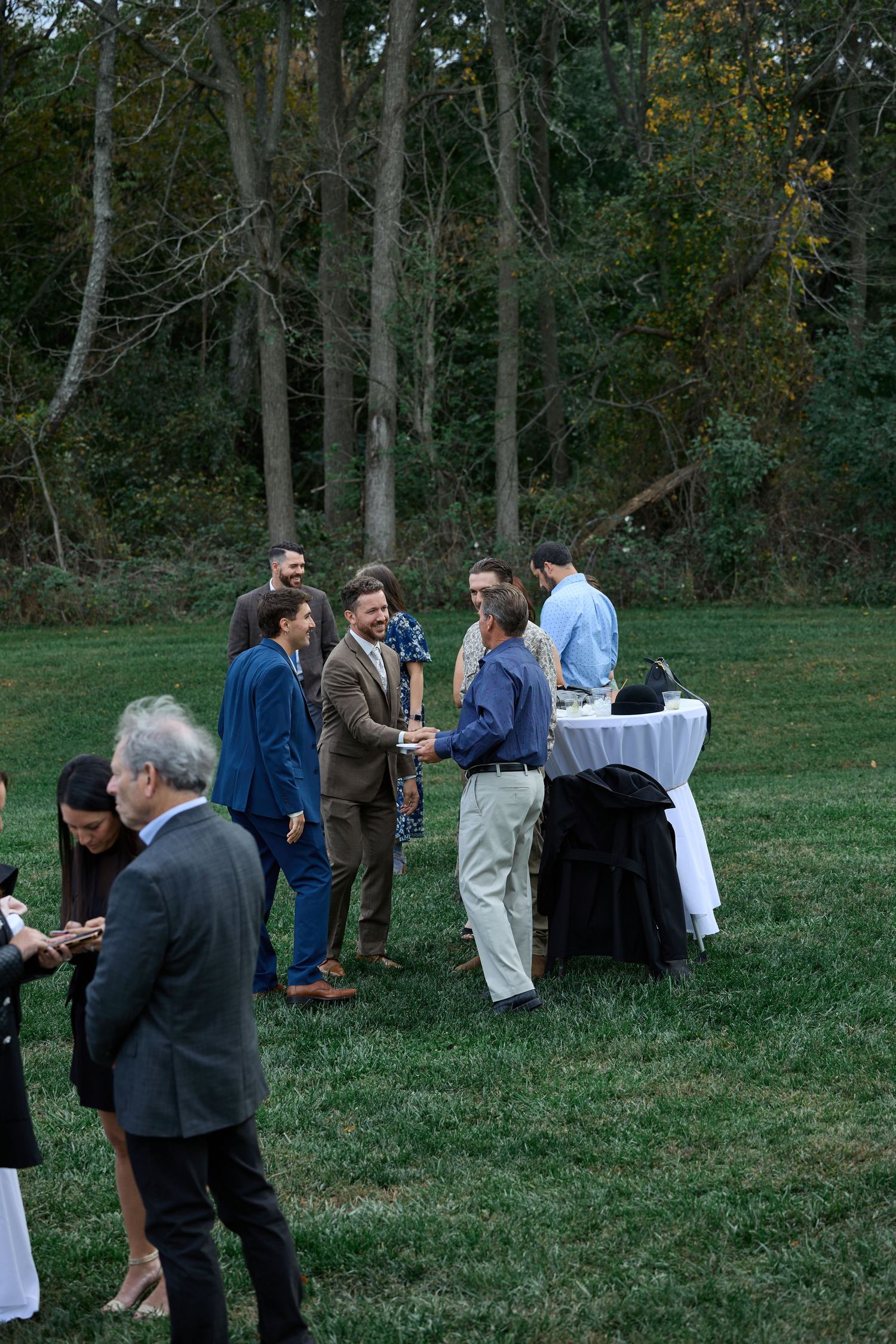 Men in suits shake hands and chat beside a cocktail table on a grassy lawn with tall trees at an outdoor wedding gathering.
