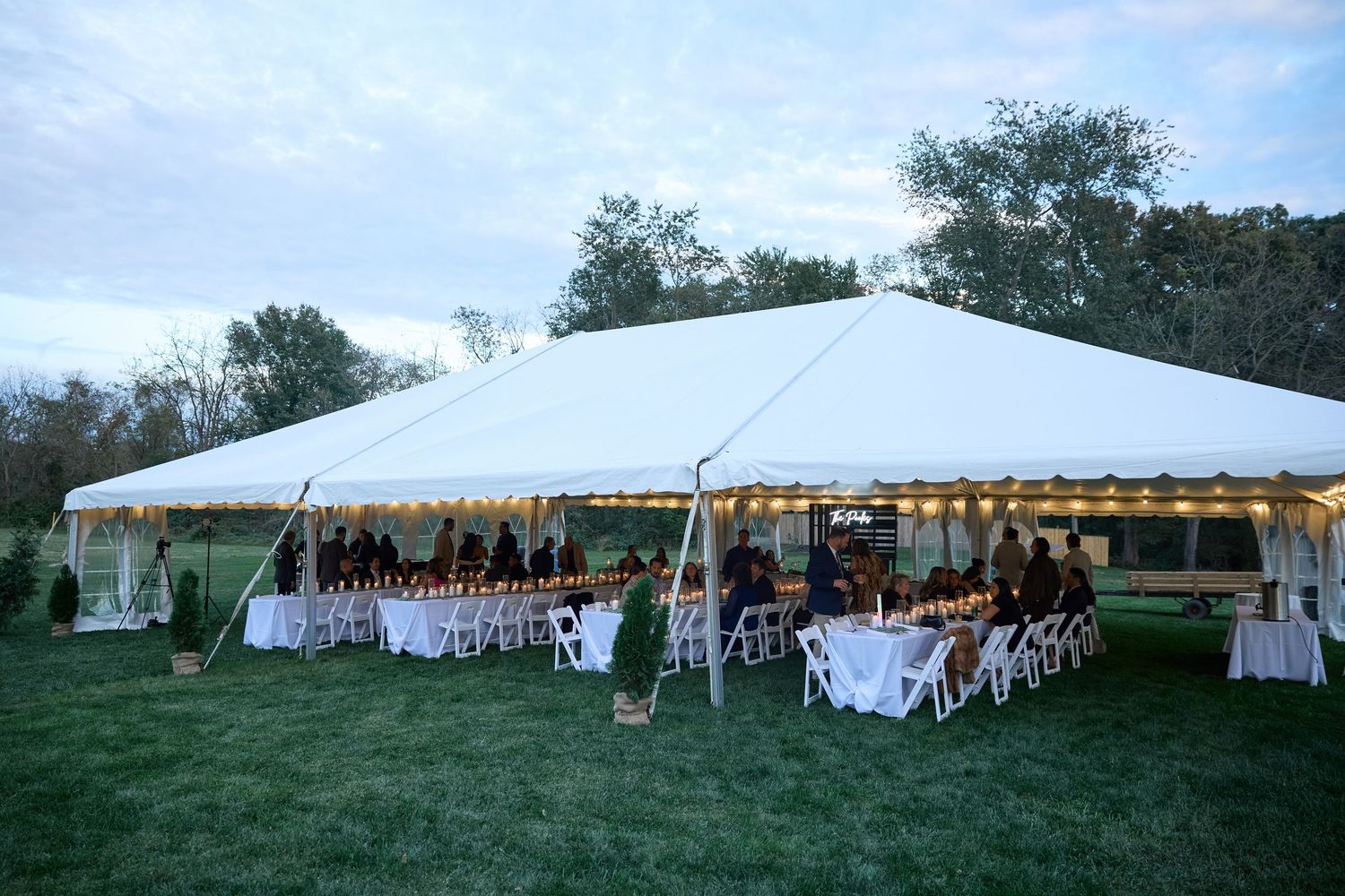 Large white tent with string lights and set banquet tables on a grassy lawn at dusk, prepared for an outdoor wedding reception.