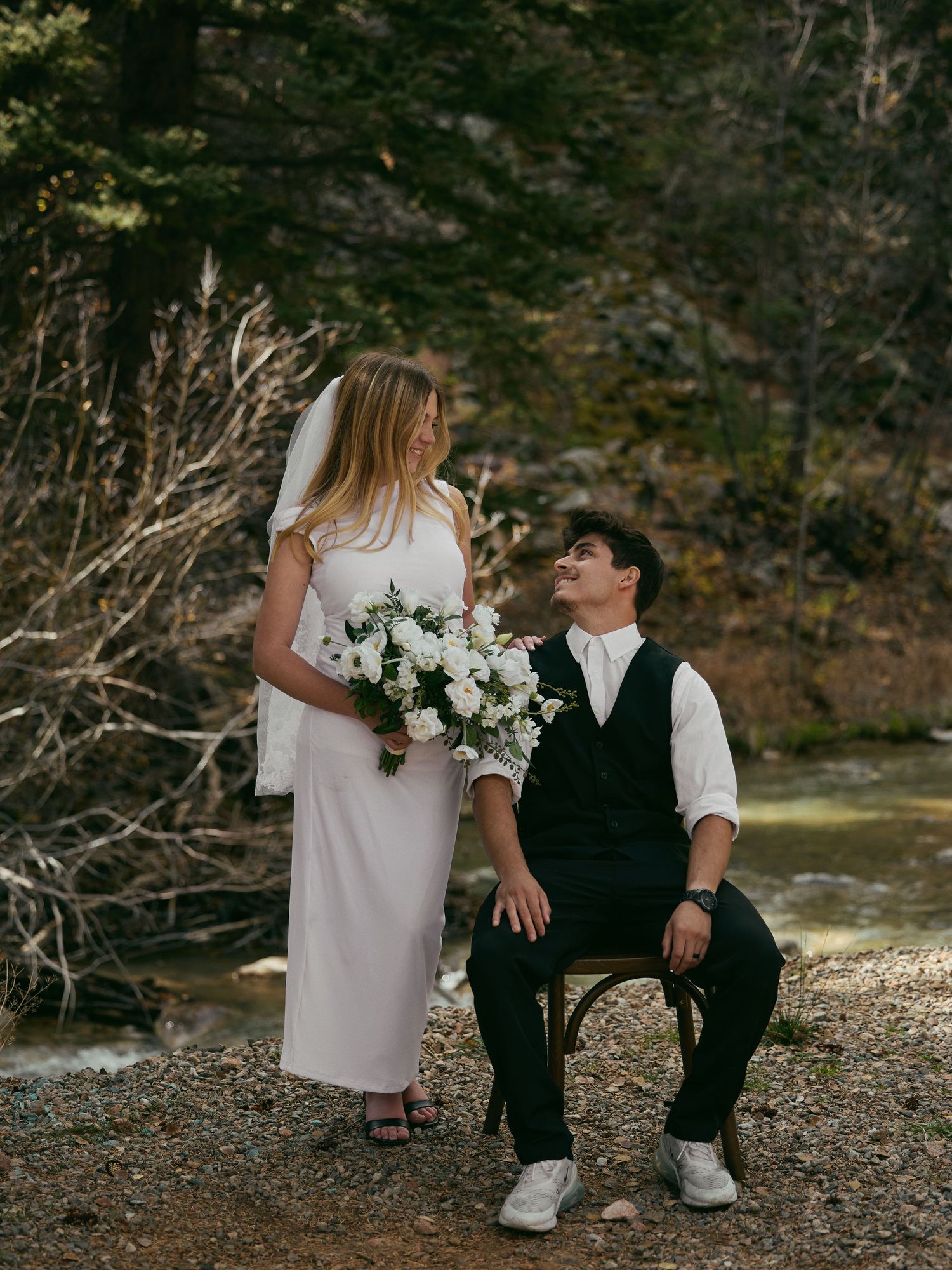 Bride in a white dress and veil holding a white bouquet smiles at a groom seated on a wooden chair beside a mountain stream.