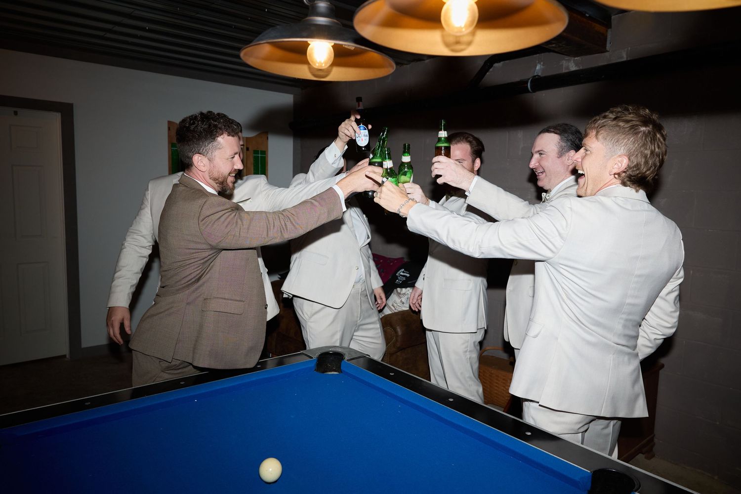 Groom and groomsmen clink green beer bottles in a toast over a blue pool table in a dimly lit room.