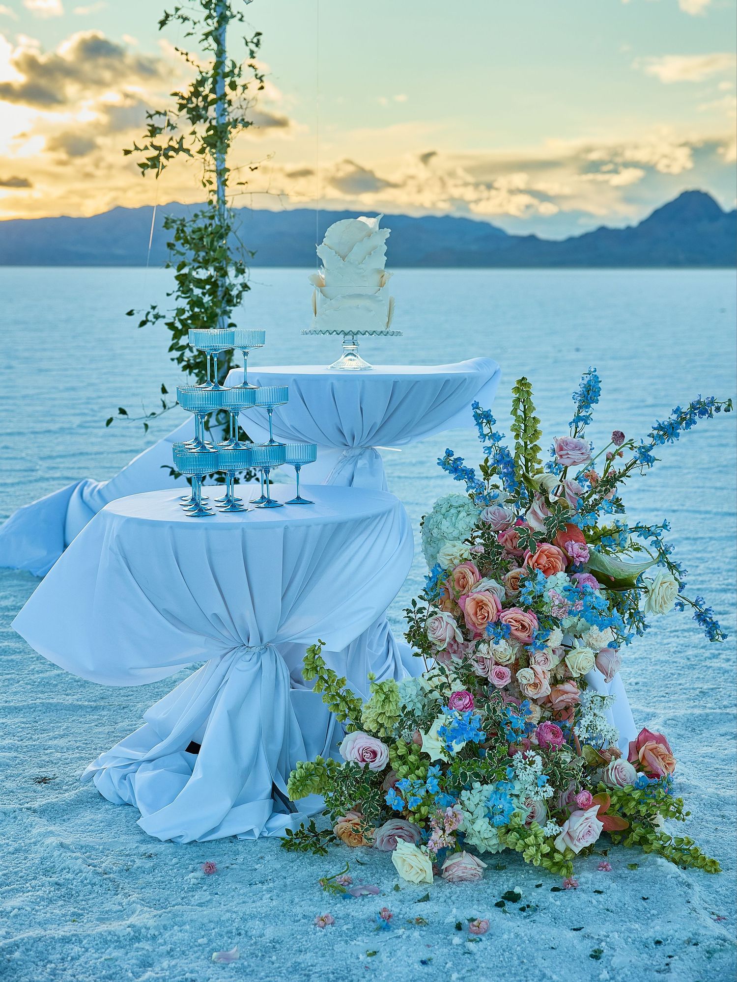 Pastel floral arrangement, champagne coupe tower and white cake on draped tables at a sunset salt flat wedding setup.