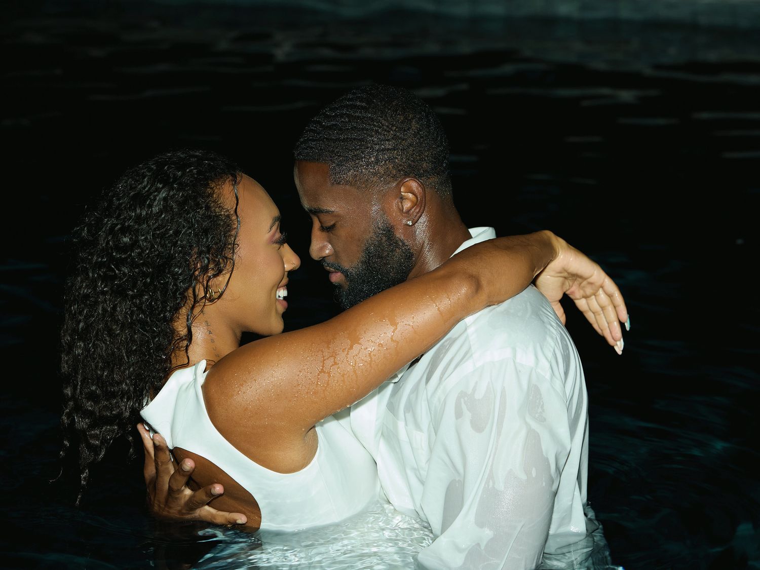 A couple in white outfits shares a tender embrace in dark pool water, capturing an intimate nighttime engagement moment.