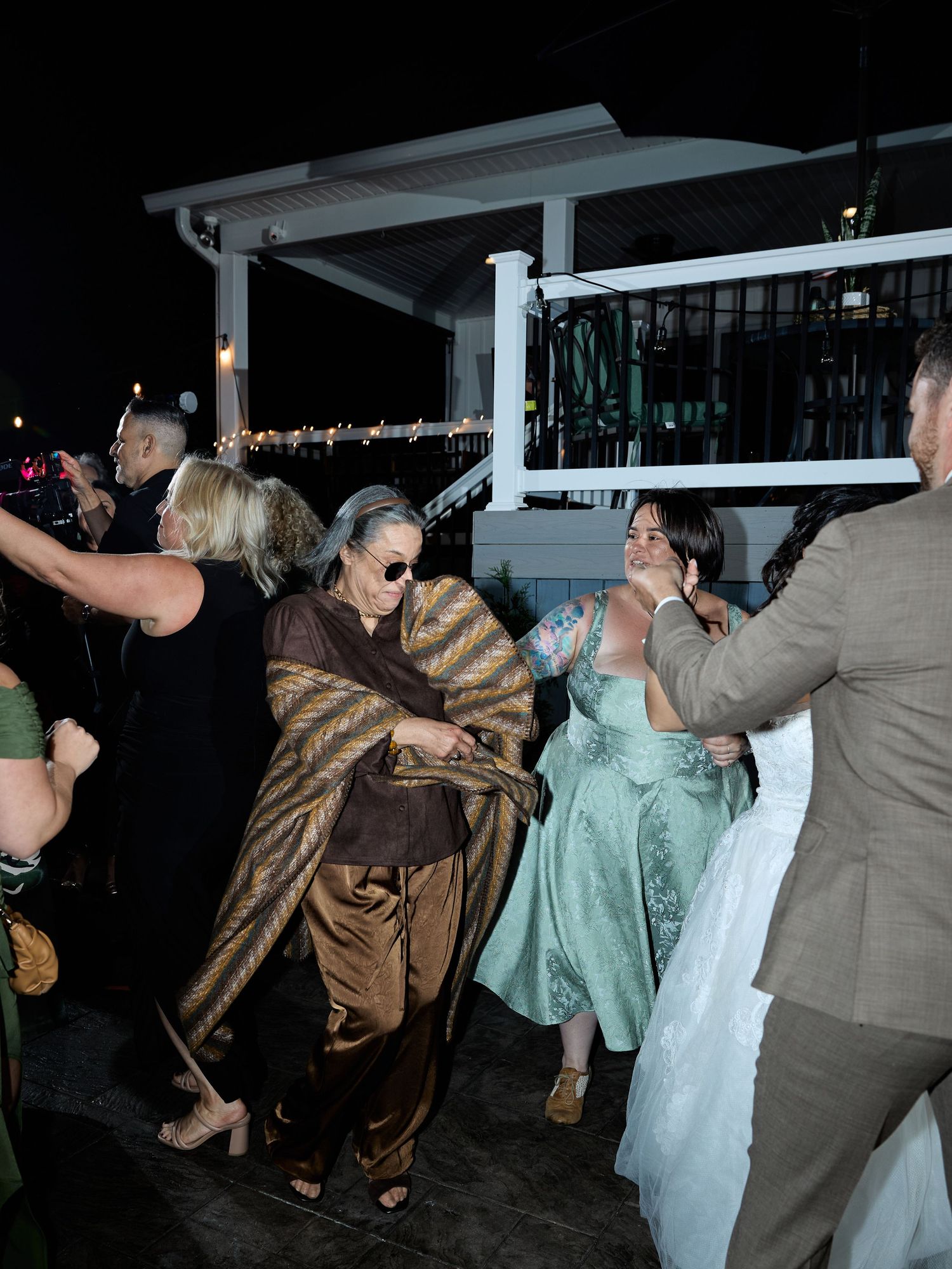 Guests dance on a night-time deck at a wedding reception, including a woman in sunglasses wrapped in a gold shawl.