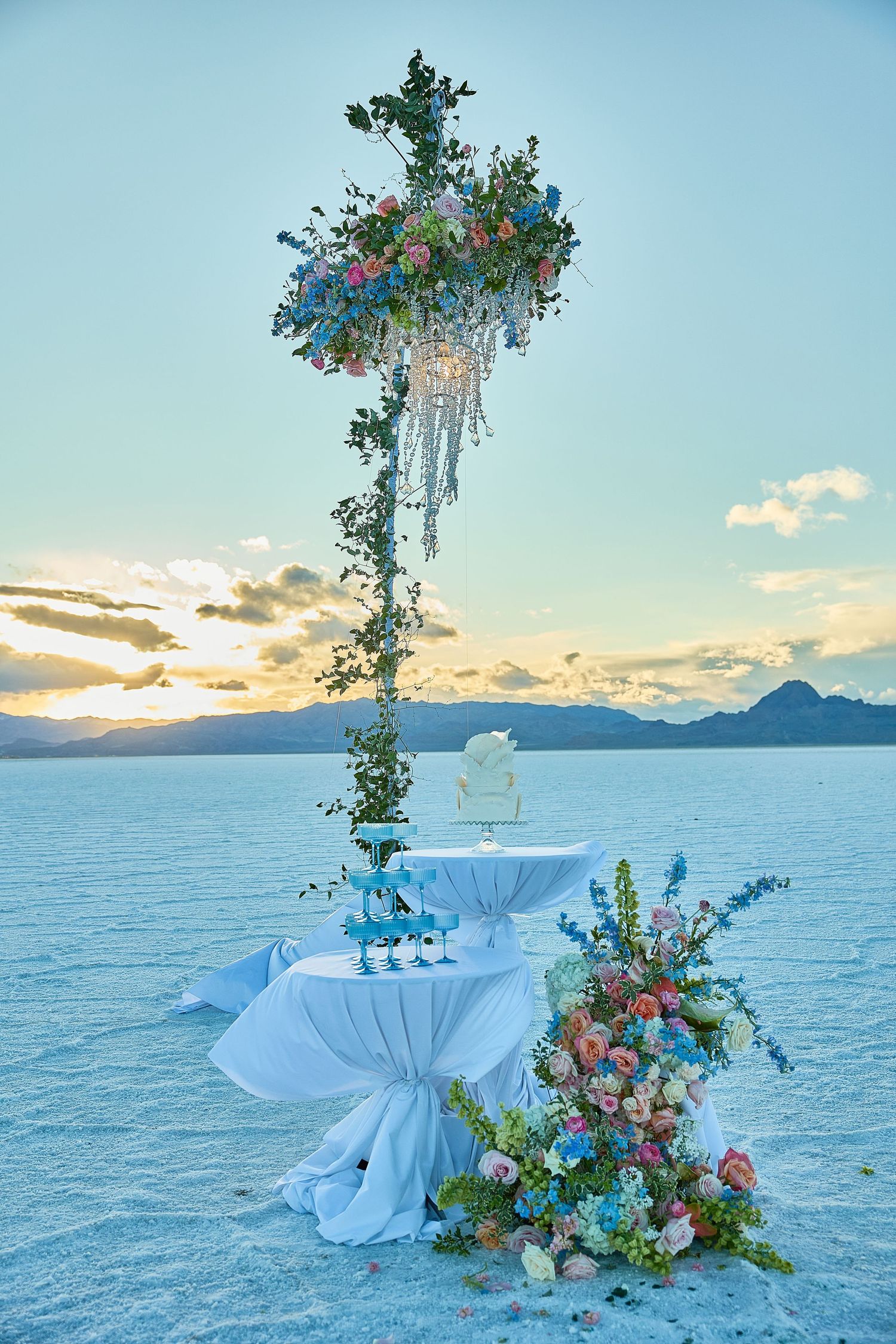 Hanging floral chandelier installation above a champagne tower, cake and flowers on a salt flat with a golden sunset sky.