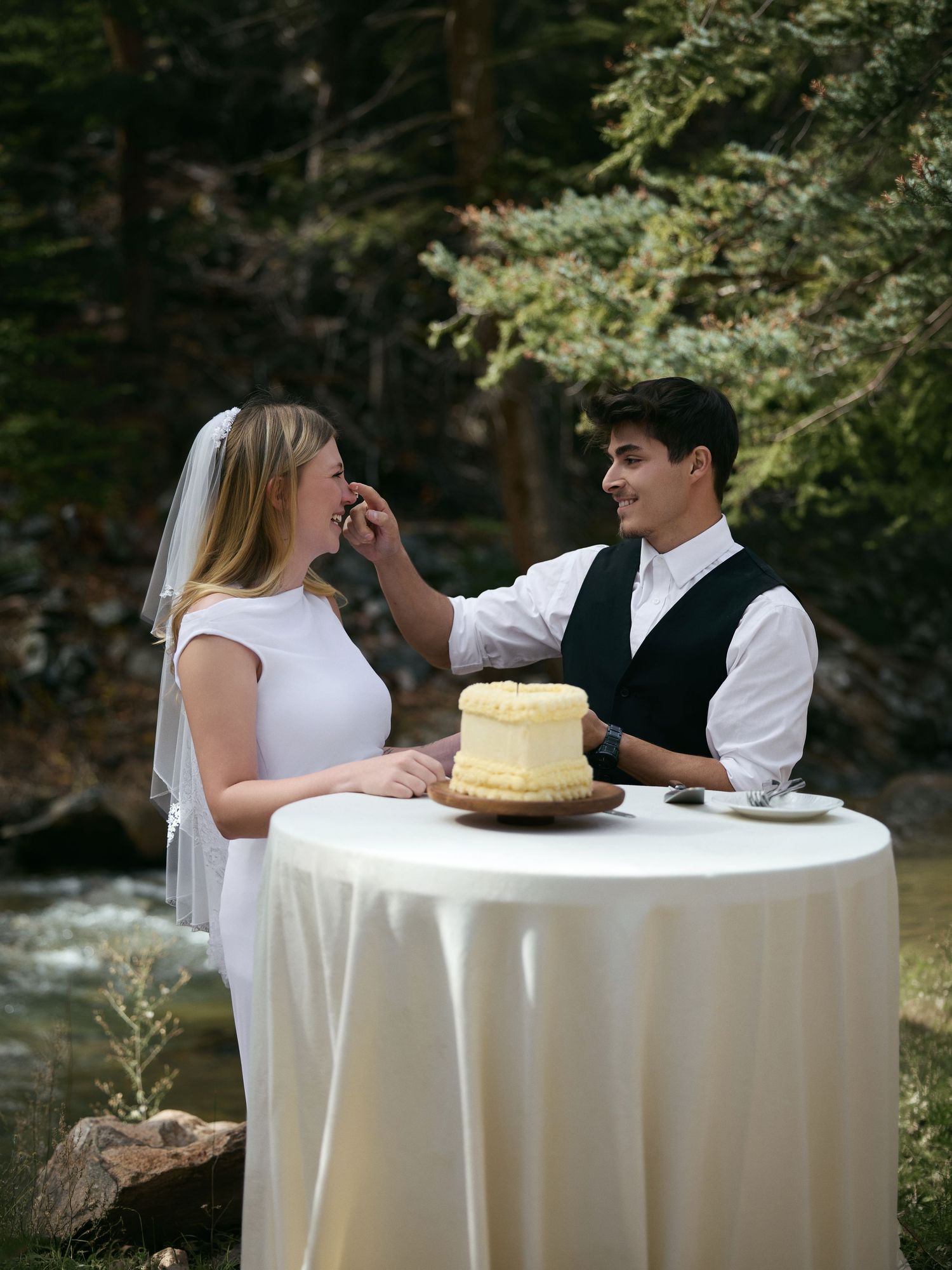 Bride and groom playfully share a bite of their small wedding cake at a draped table by a forest stream.