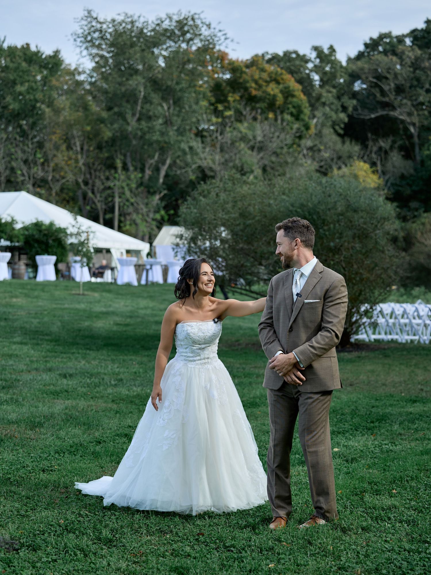 Bride in a strapless white gown reaches out to the groom in a brown suit on a lawn near white chairs and a tent.