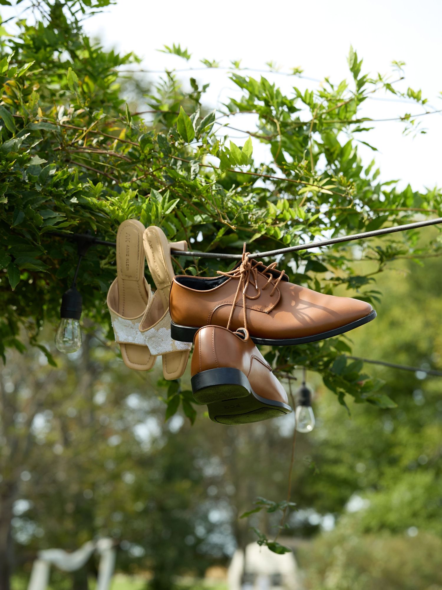 Brown leather groom's shoes and cream women's heeled sandals hanging from a string of café lights among green leaves.