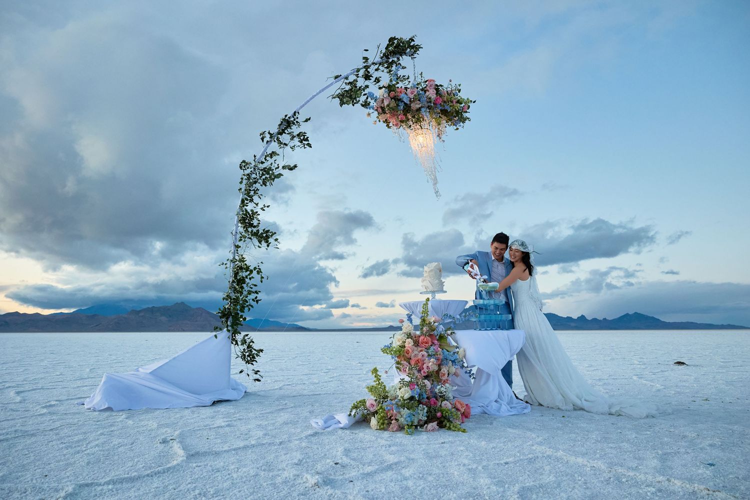 Bride and groom share a tender moment by a floral arch and bouquet display on a white salt flat beneath stormy skies.