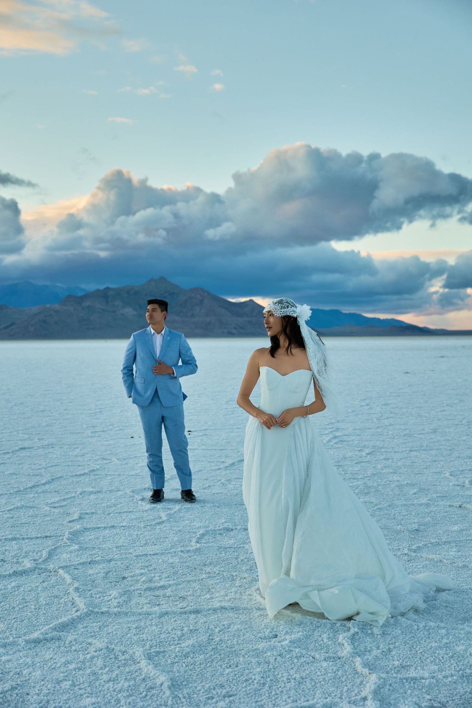Bride in a strapless white gown and veil stands in front of her groom in a blue suit on a salt flat with cloudy mountain views.