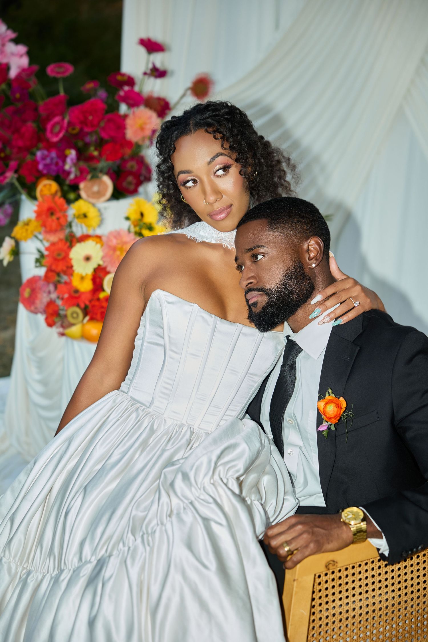 A bride rests against her seated groom in a black suit, with a bright floral arrangement and white draping behind them.