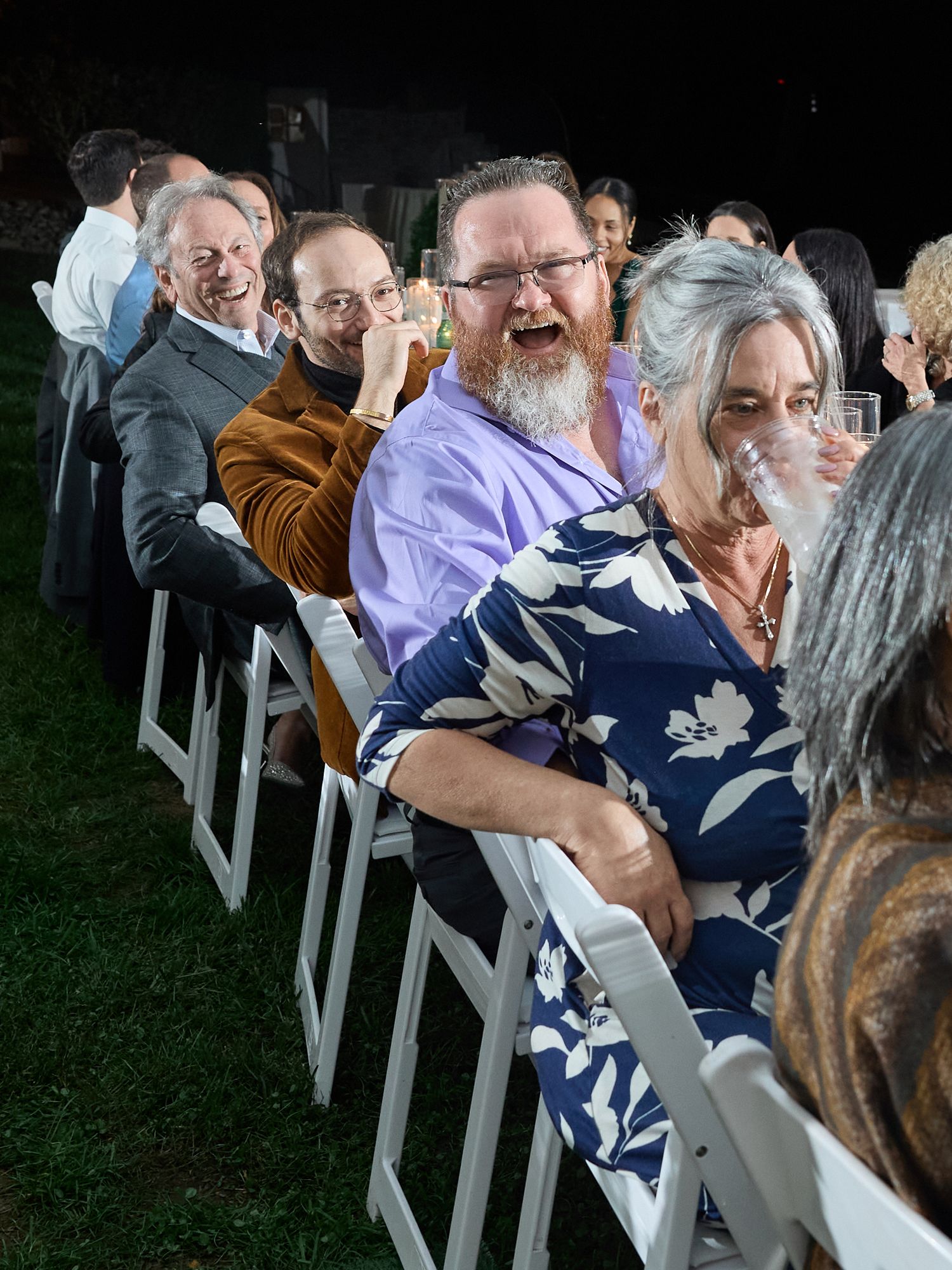 Wedding guests seated in white folding chairs on the lawn laugh heartily during an evening reception speech.