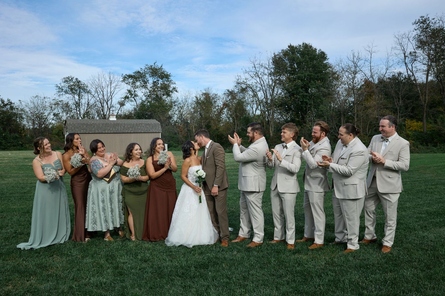 Bridesmaids in earthy dresses and groomsmen in tan suits applaud as the bride and groom kiss on a grassy lawn.