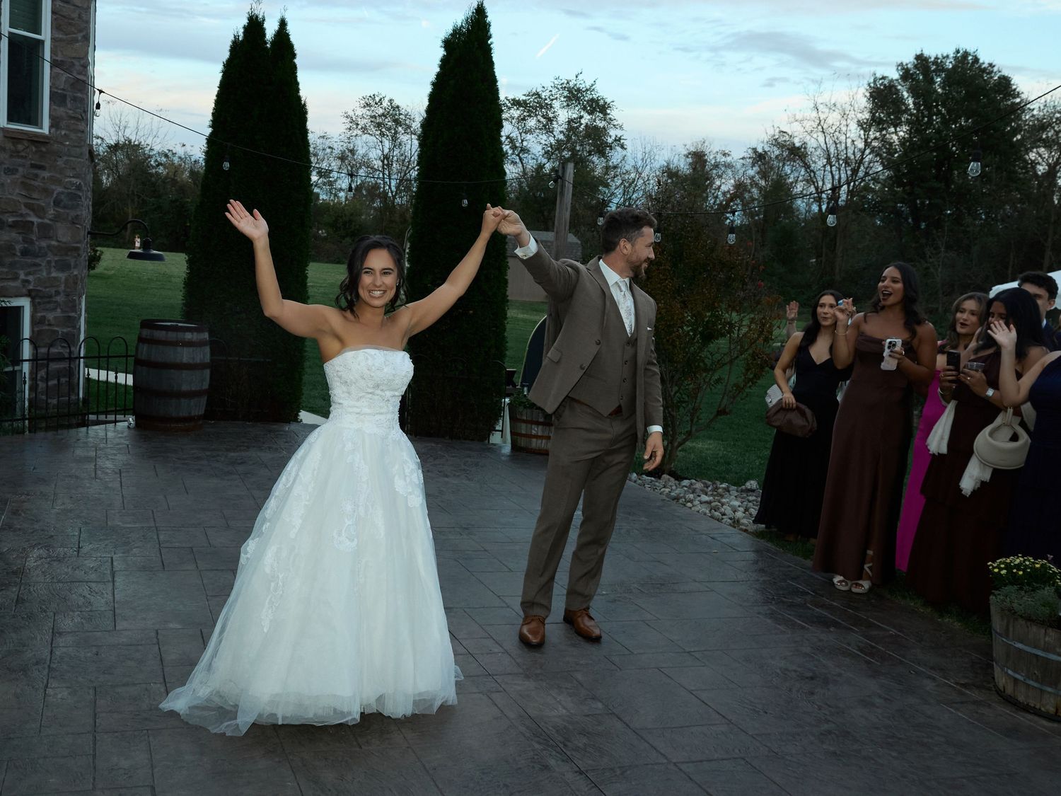 A bride in a strapless white gown raises her arm and twirls with her groom on a patio as guests watch at dusk.