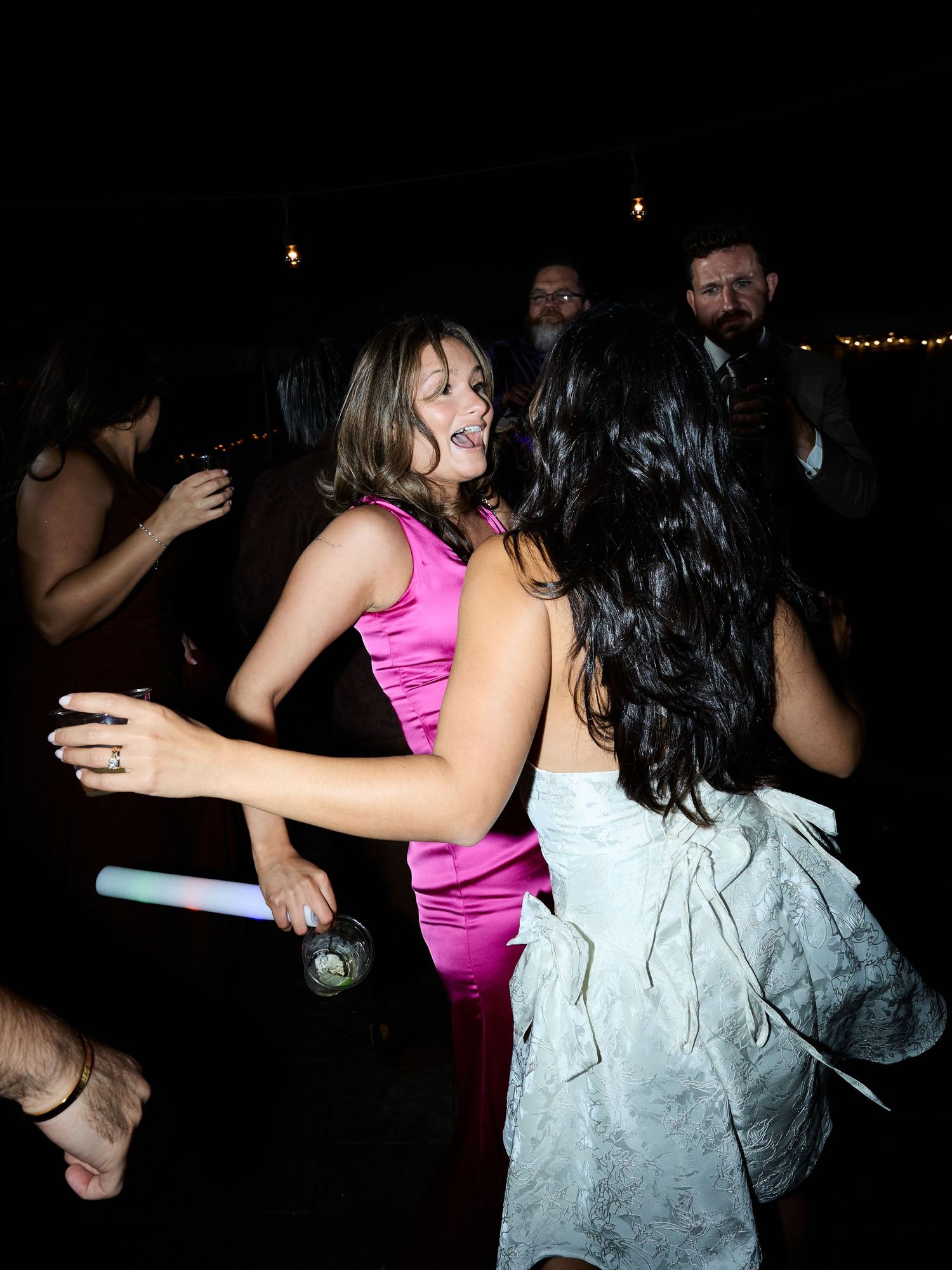 Two women in pink and pale blue dresses dance together at a night wedding reception with glow sticks nearby.