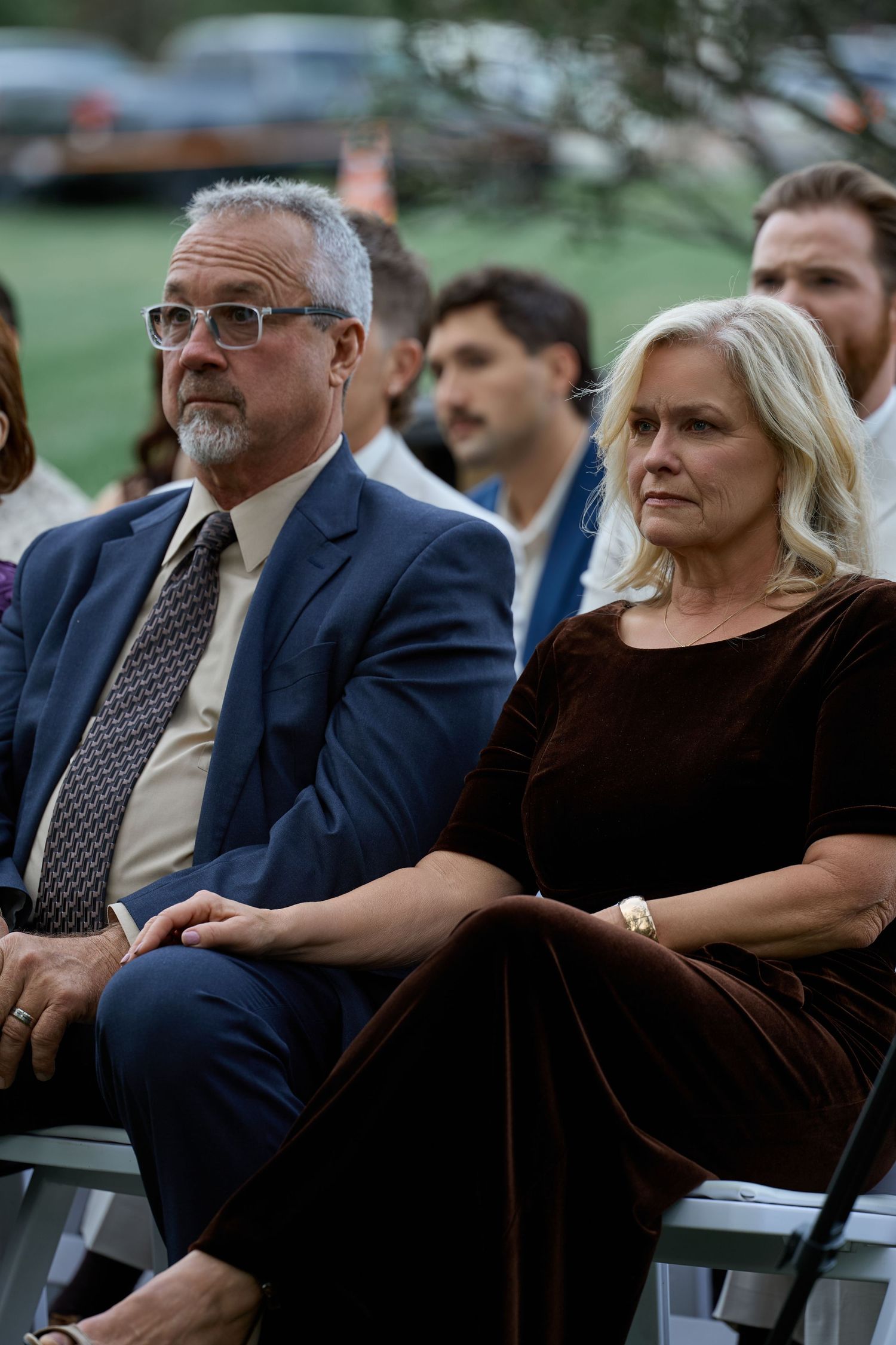 Two wedding guests, a gray-haired man in glasses and a blue suit and a woman in a velvet dress, sit attentively during an outdoor ceremony.