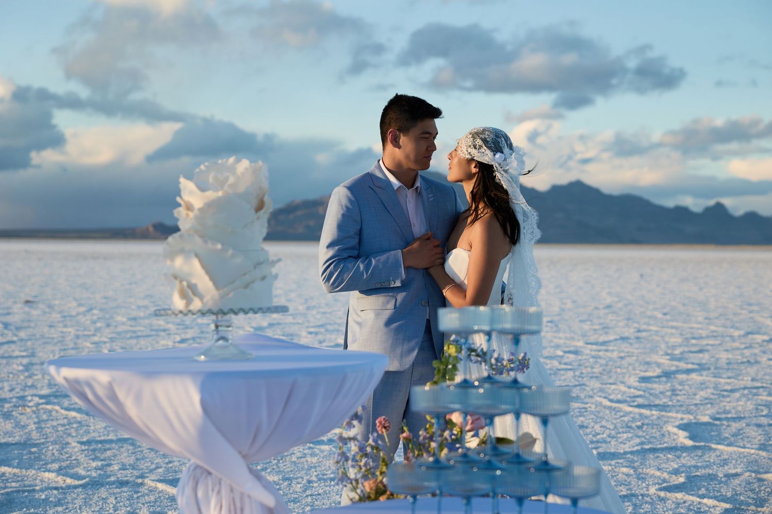 Groom in a blue suit holds his veiled bride beside a table with a champagne tower on a glistening salt flat.