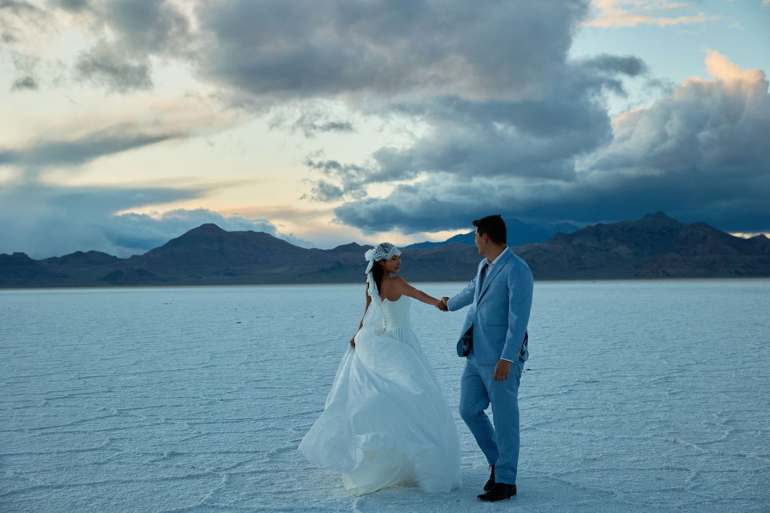 Bride in a long white gown and veil walks hand in hand with the groom in a blue suit across a salt flat at dusk.
