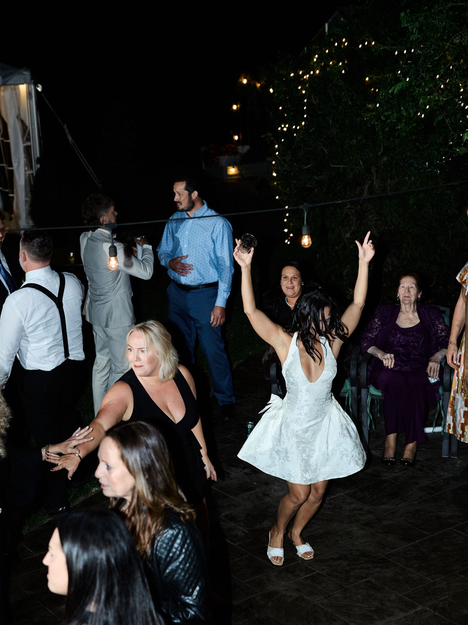 Wedding guests dance outdoors at night under string lights while a woman in a white dress raises her arms.