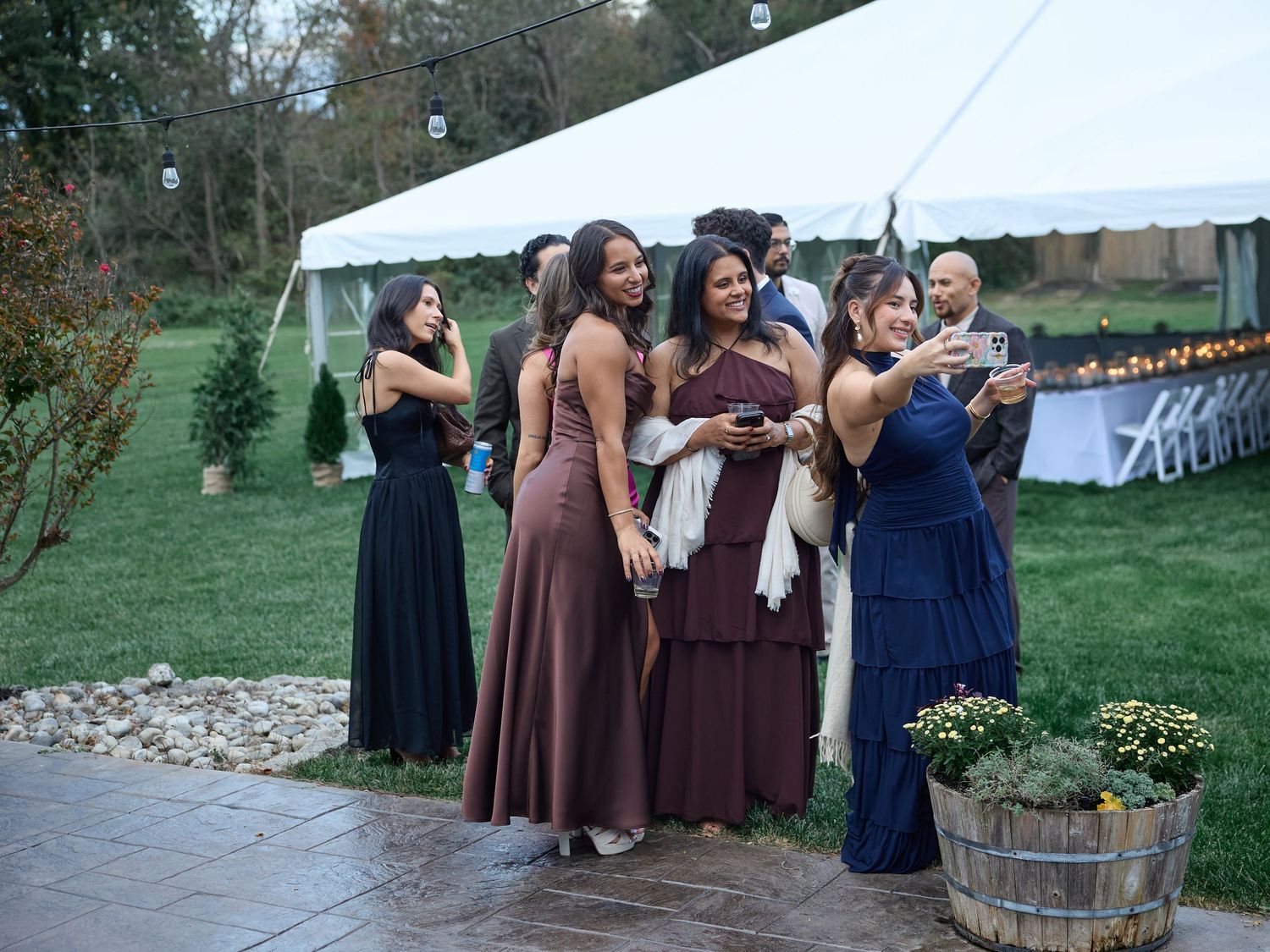 Bridesmaids and guests in elegant dresses pose for a group selfie on a stone patio beside a white wedding tent.