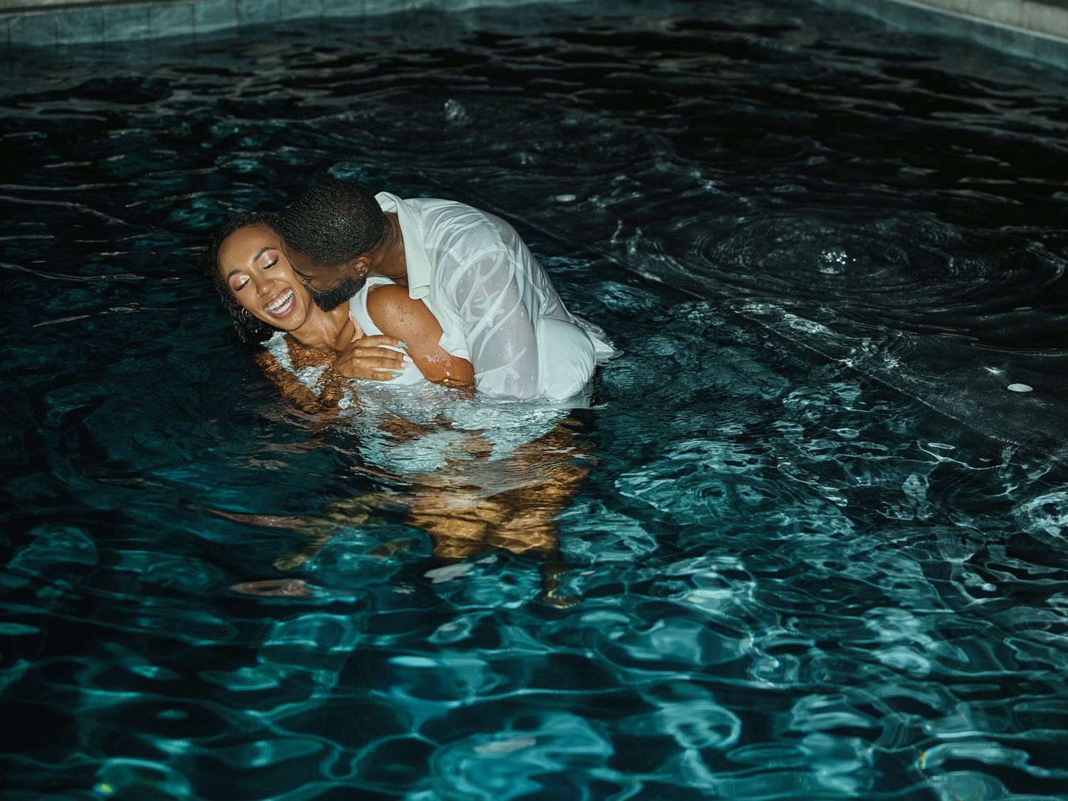 A laughing woman in white is held by a man in a white shirt in the dark rippling water of a pool.