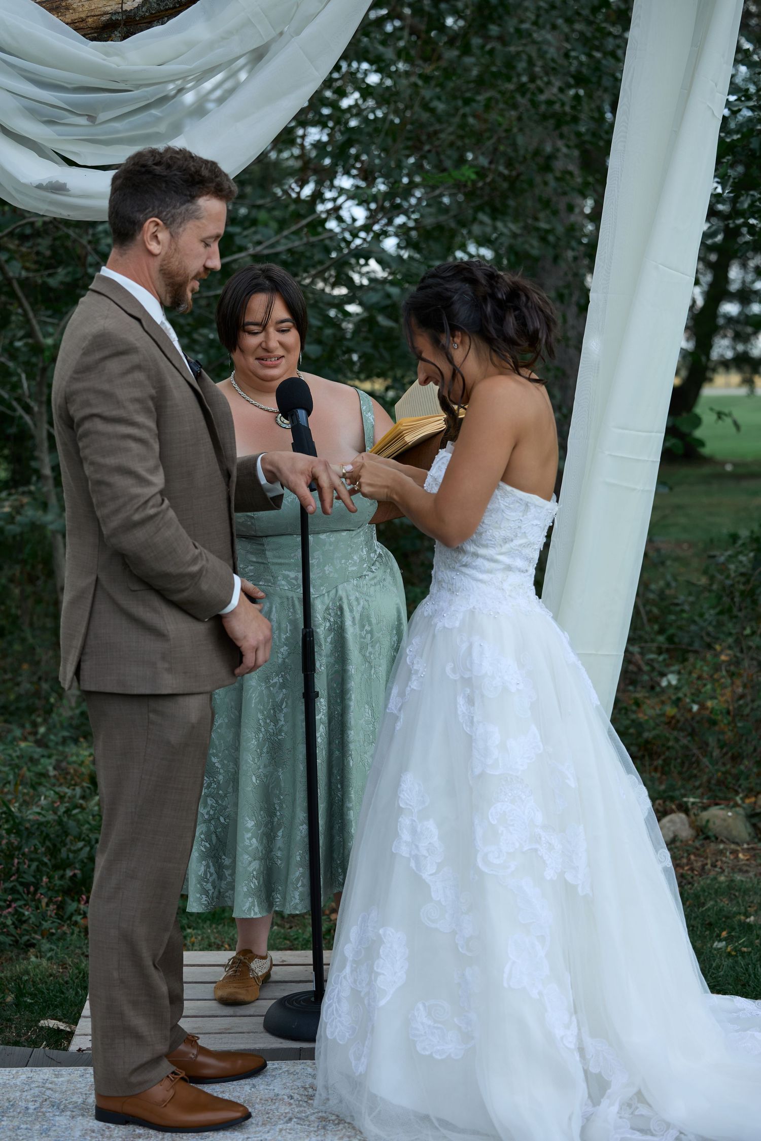 The bride in a white lace gown places a ring on the groom's finger as the officiant stands behind them under a fabric canopy.