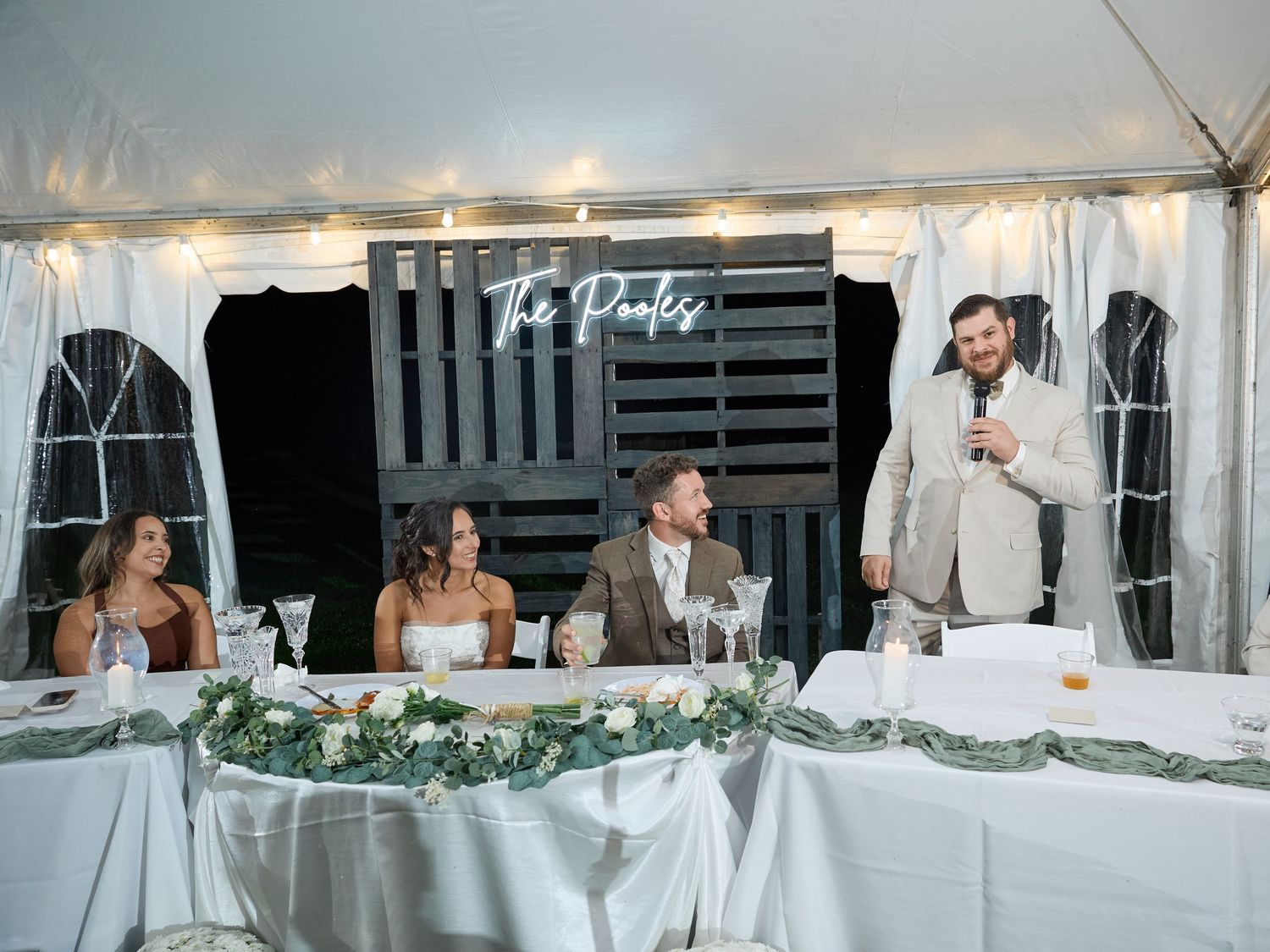 Groomsman gives a toast with a microphone as the bride, groom and bridesmaid sit at the floral-decorated head table with a neon Pooles sign.
