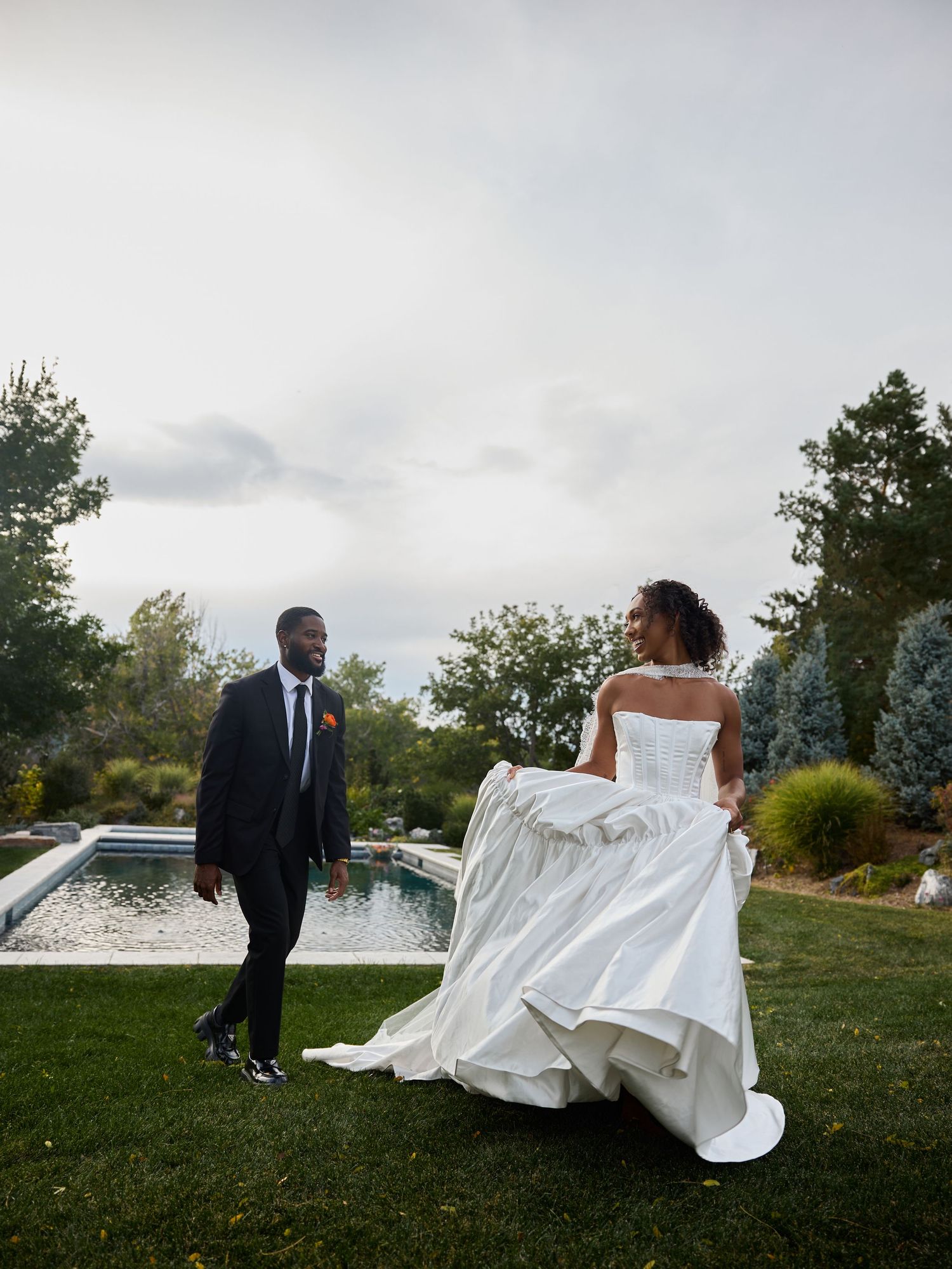 Groom in a dark suit walks across the grass as the bride lifts her voluminous white gown beside a garden pool.
