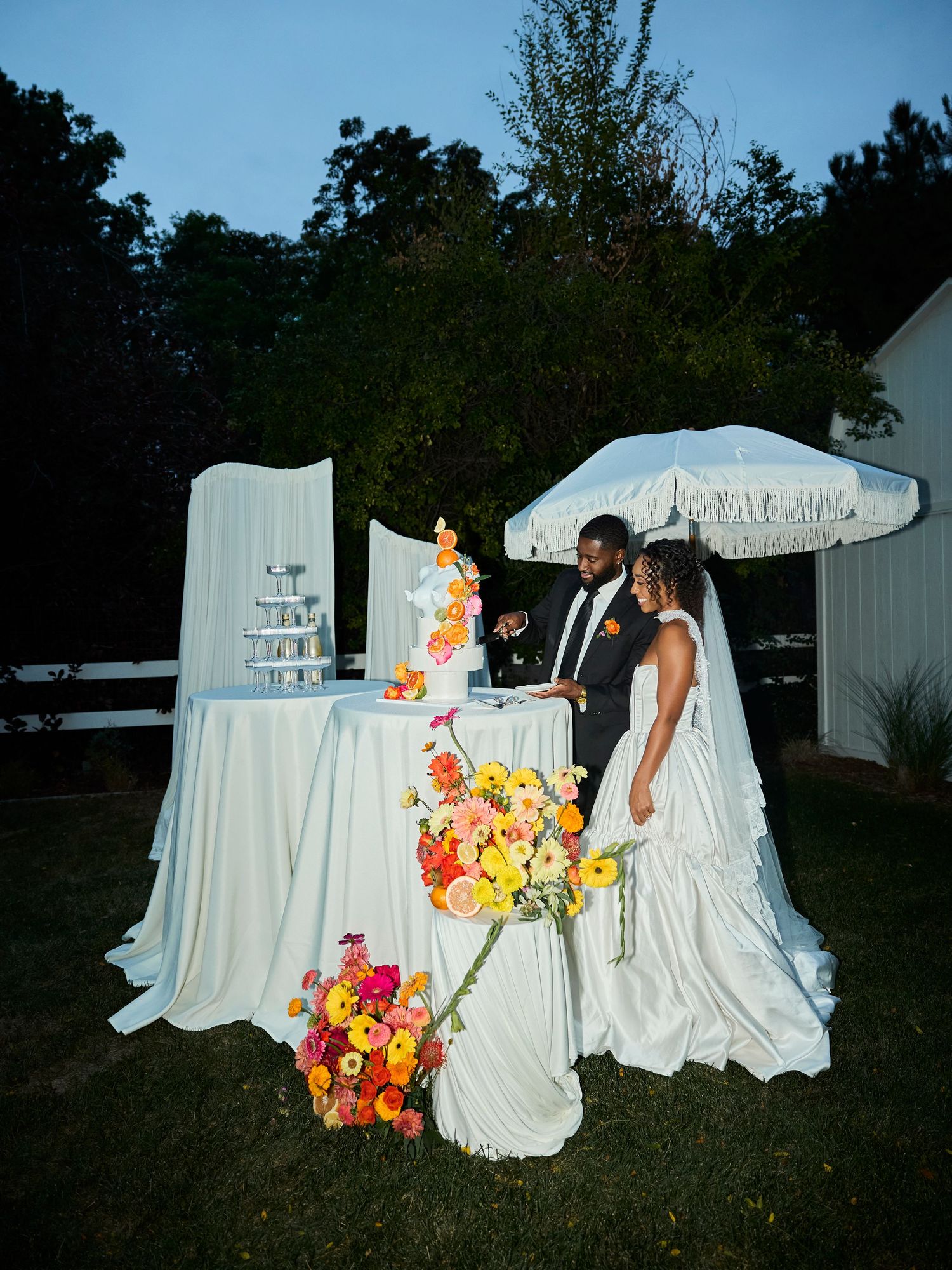 A bride and groom cut a tiered wedding cake adorned with bright orange and pink flowers outdoors at dusk beneath a fringed white umbrella.