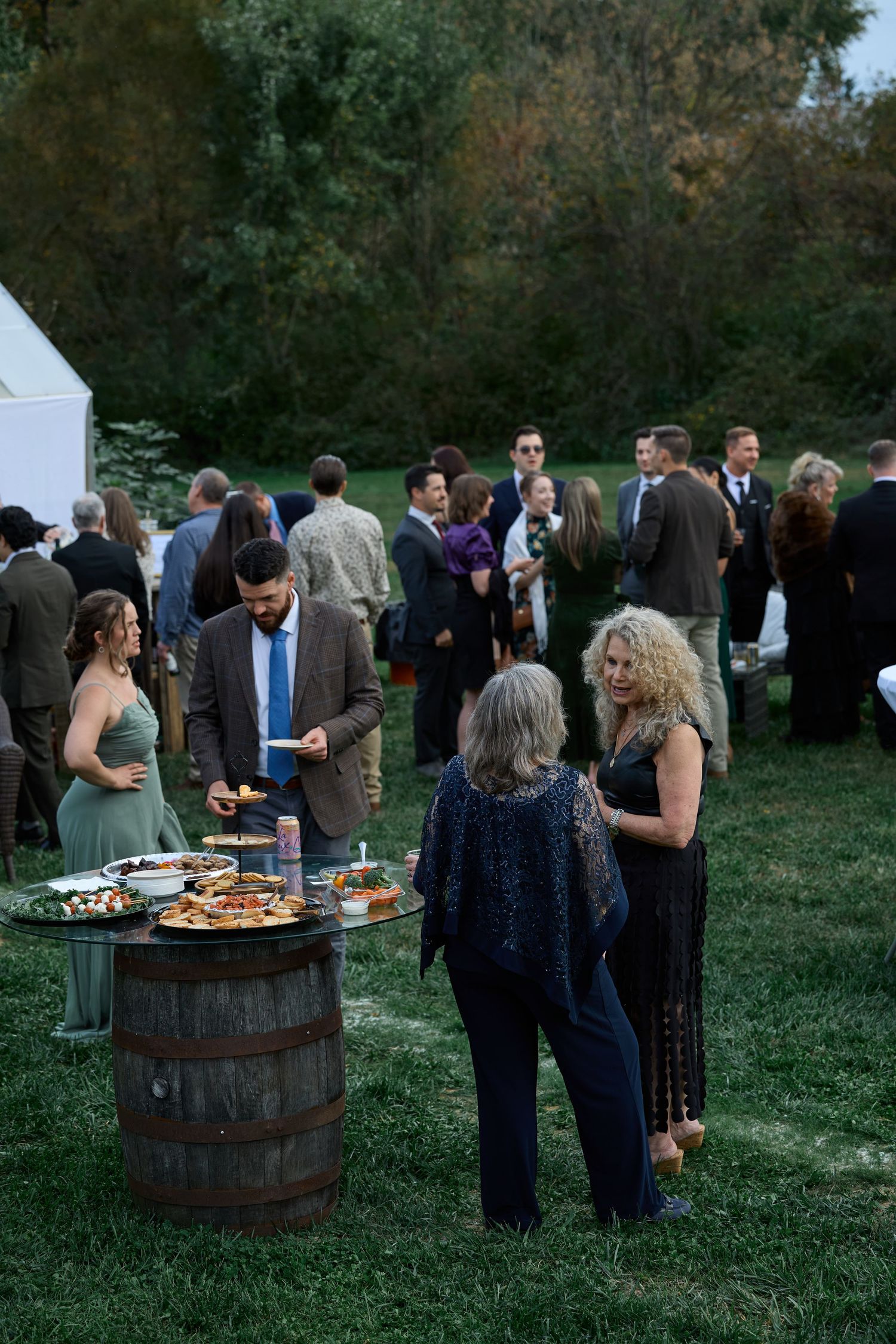 Guests in dressy attire gather around a whiskey barrel table with appetizers on a grassy lawn during an evening wedding cocktail hour.